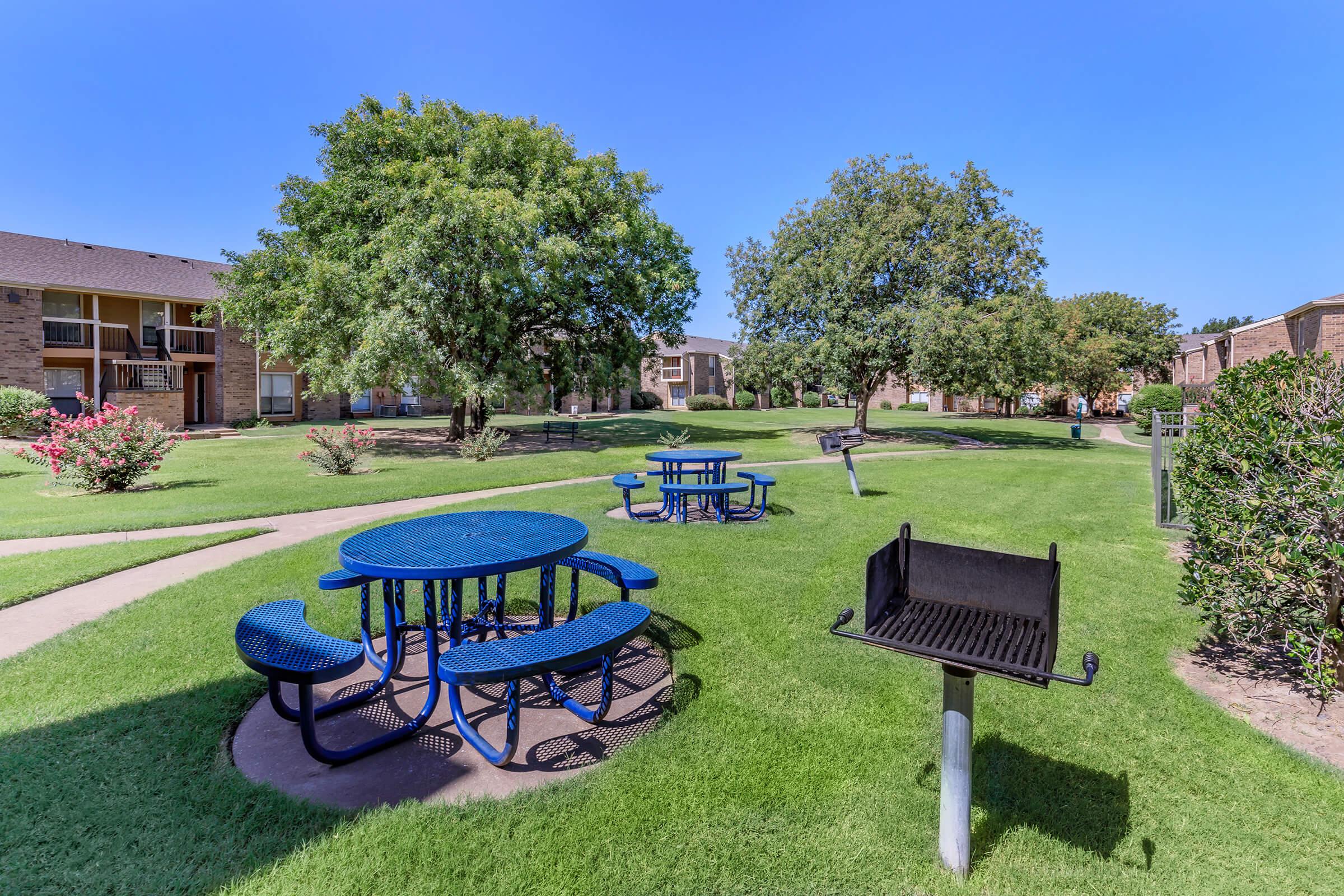 A sunny outdoor area featuring blue picnic tables and a charcoal grill, surrounded by well-maintained grass and trees. The scene includes a walking path, with several buildings visible in the background, creating a pleasant and inviting communal space.
