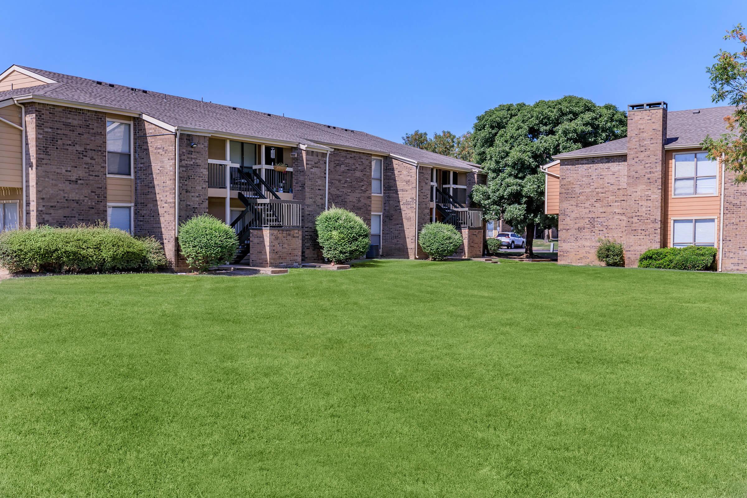 A sunny view of a residential apartment complex featuring two buildings made of brick, surrounded by well-manicured lawns and shrubs. The clear blue sky adds to the bright atmosphere of the outdoor space.