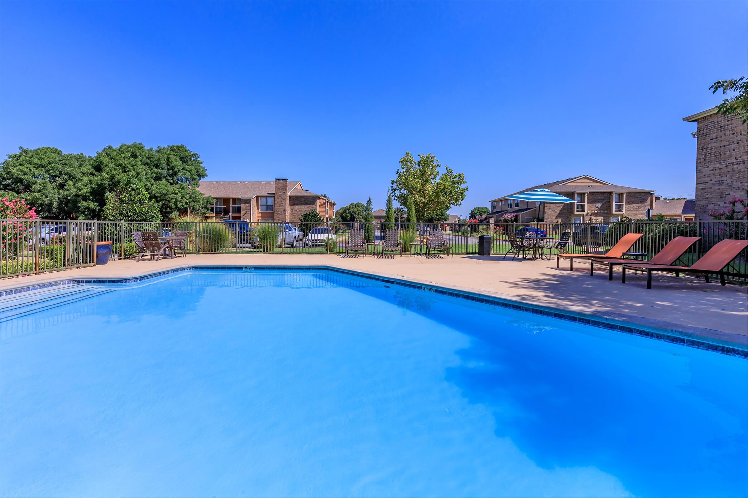 A clear blue swimming pool surrounded by lounge chairs and lush greenery. In the background, there are residential buildings and trees under a bright blue sky, creating a serene outdoor atmosphere perfect for relaxation.