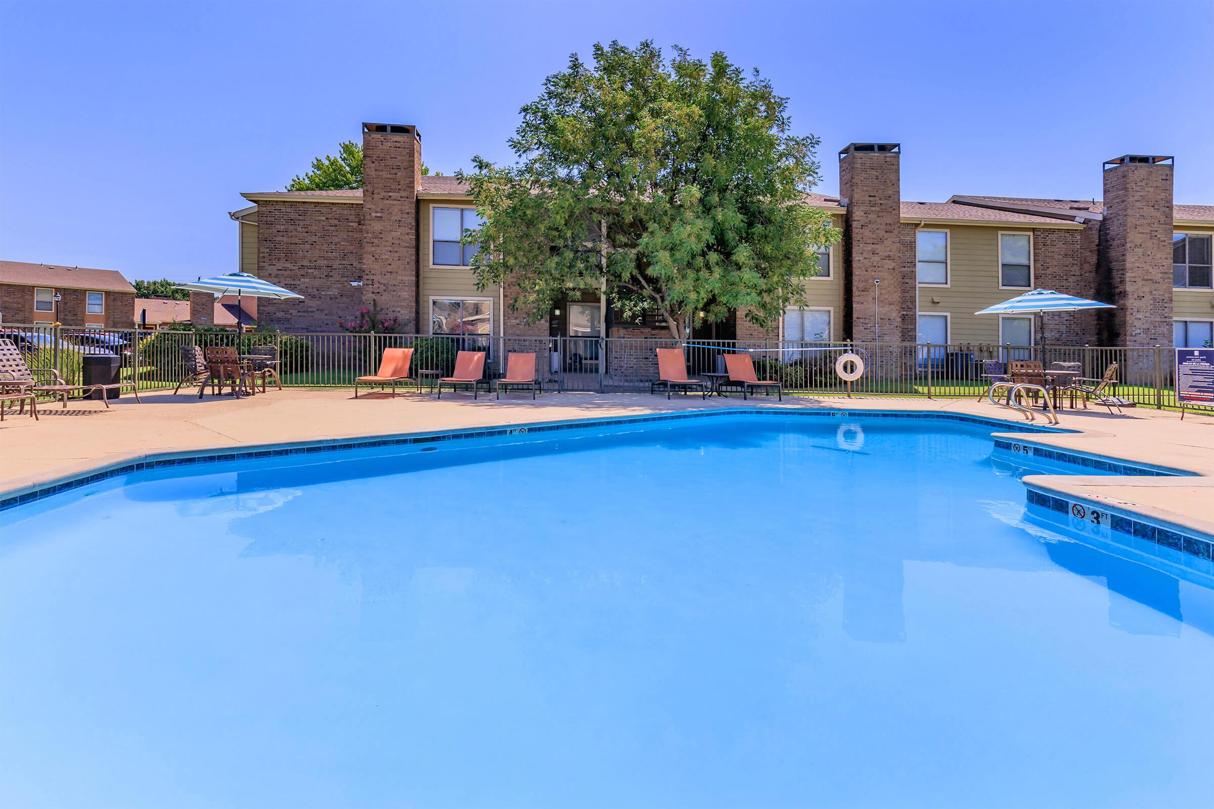 A clear blue swimming pool surrounded by a paved area, with lounge chairs and umbrellas set up for relaxation. In the background, a two-story apartment building with greenery and trees provides a pleasant setting. The sky is bright and sunny, creating a tranquil atmosphere.