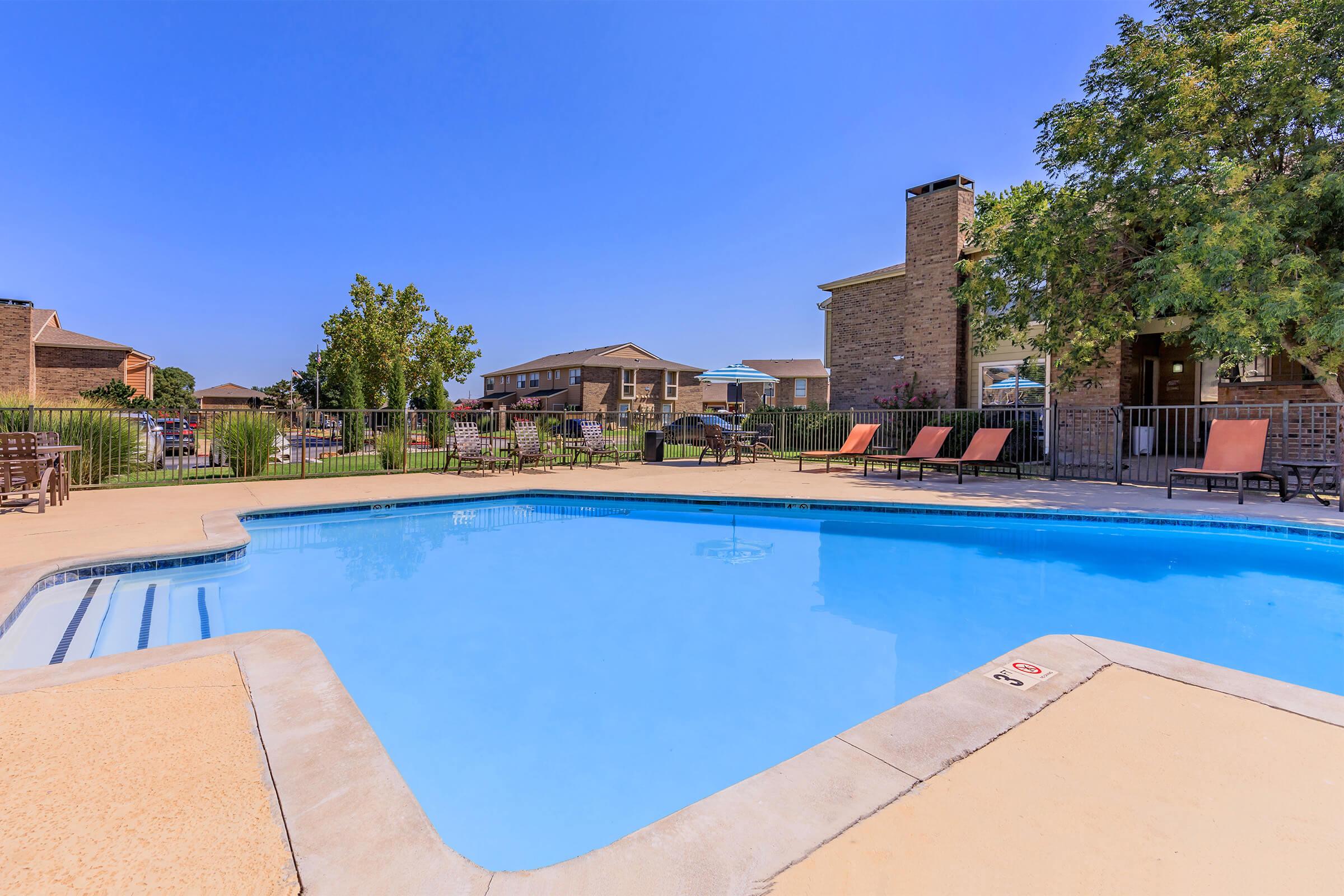 A clear blue swimming pool surrounded by a concrete deck, lounge chairs, and a grassy area. In the background, there are several buildings and trees under a bright blue sky, indicating a sunny day.