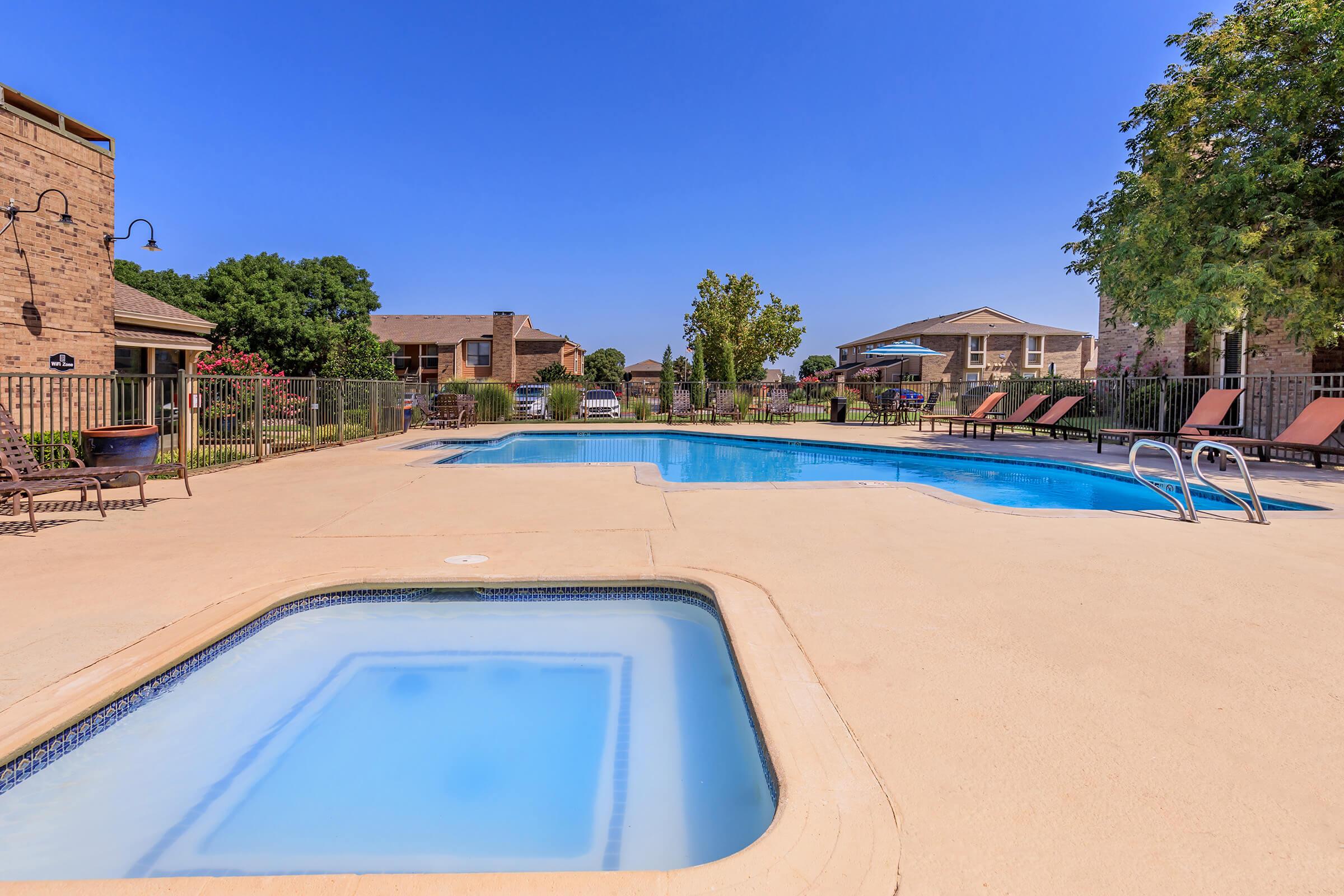 A sunny outdoor swimming pool area featuring two pools: a larger rectangular pool and a smaller hot tub. Surrounding the pools are lounge chairs and a fence, with green trees and buildings visible in the background under a clear blue sky.
