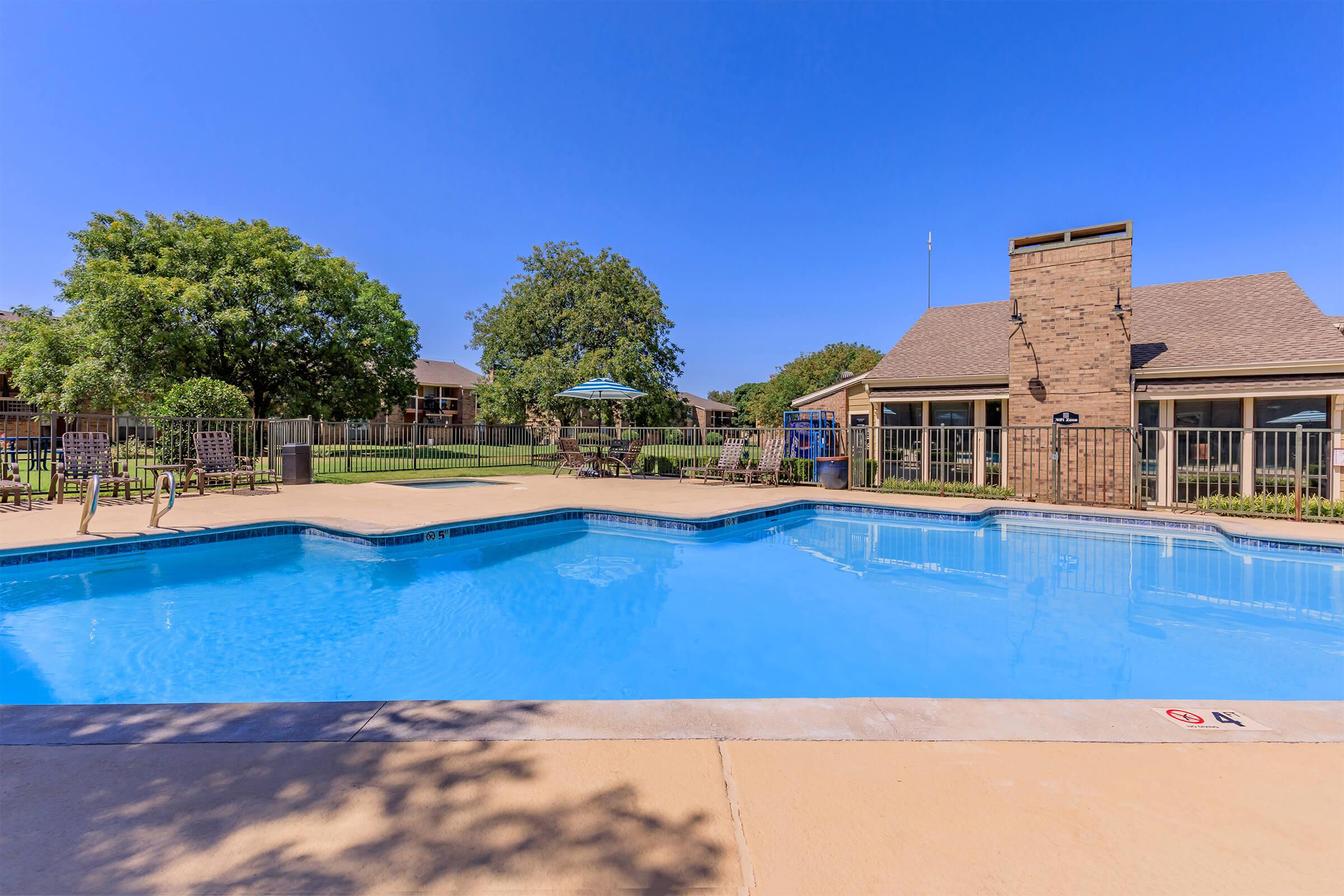A clear blue swimming pool surrounded by a patio area. There are lounge chairs and a shaded umbrella nearby. In the background, well-maintained grass and trees are visible, along with a building that features large windows. The sky is bright and sunny, creating a welcoming outdoor space.