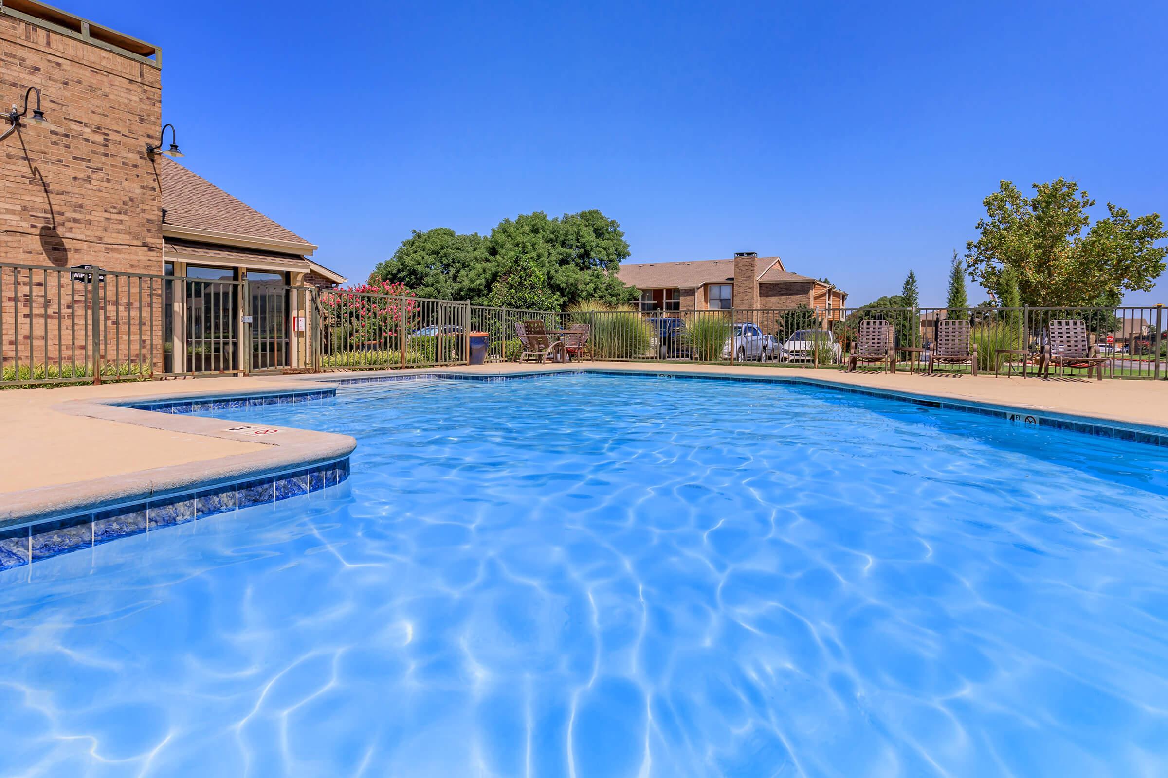 A clear blue swimming pool surrounded by a patio, with green trees and residential buildings in the background. The sky is bright blue, creating a sunny and inviting atmosphere. Laid-back seating areas are visible near the pool.
