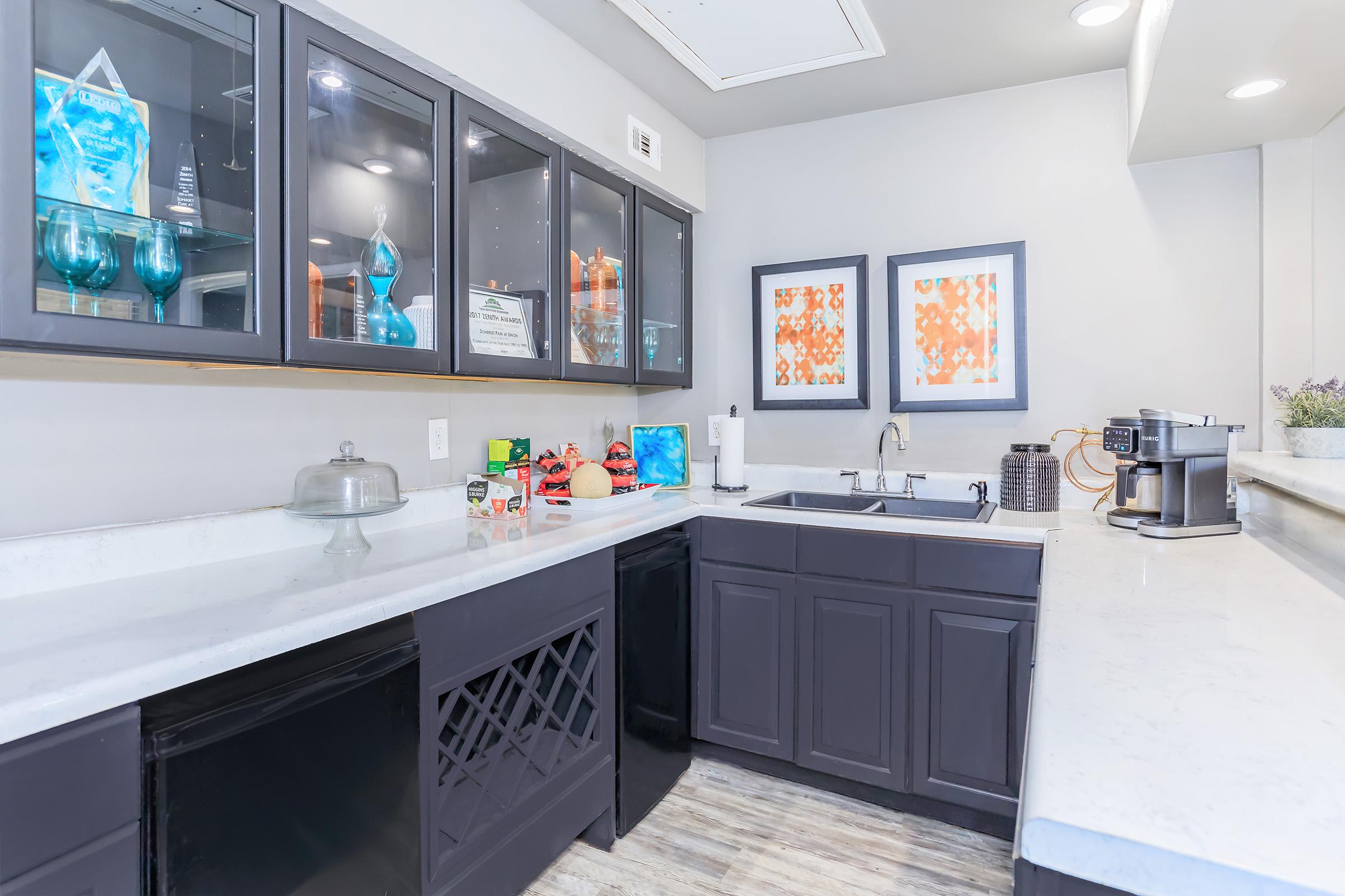 Modern kitchen featuring dark cabinetry with glass doors displaying decorative items. A white countertop includes a sink and coffee maker, complemented by framed artwork on the wall. The space is well-lit, with a clean, organized layout and a touch of color from kitchen accessories.