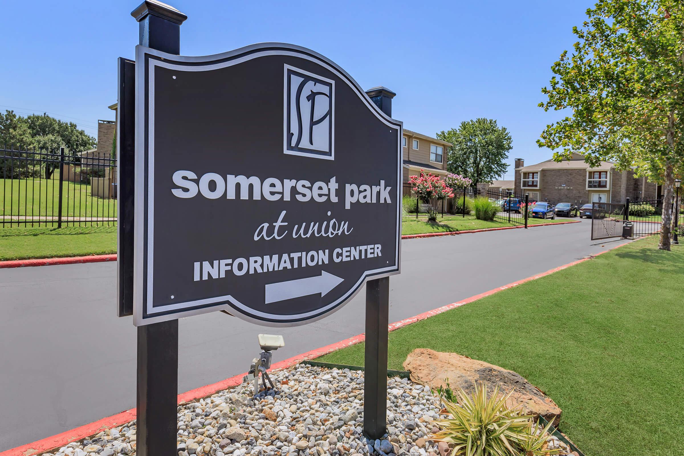 Sign for Somerset Park at Union, indicating the location of the Information Center. The sign features a decorative design and is situated near well-maintained landscaping, with grass and rocks, on a clear blue sky day. The road leading to the center is paved and lined with trees and flowering plants.