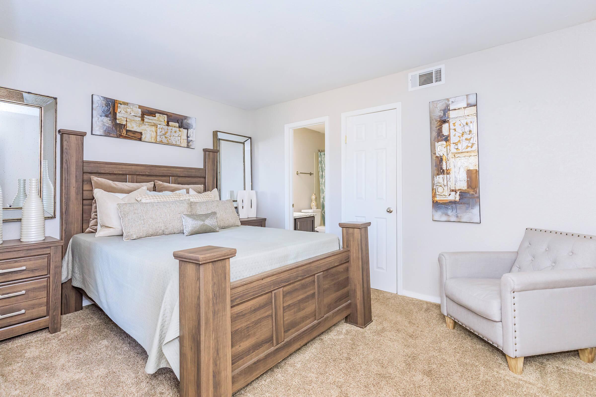 A cozy bedroom featuring a wooden bed frame with decorative pillows, a beige and gold color scheme, and matching nightstands. The room has soft carpet, a gray armchair in the corner, and wall art above the bed. A door leads to a bathroom area, providing an inviting and serene atmosphere.