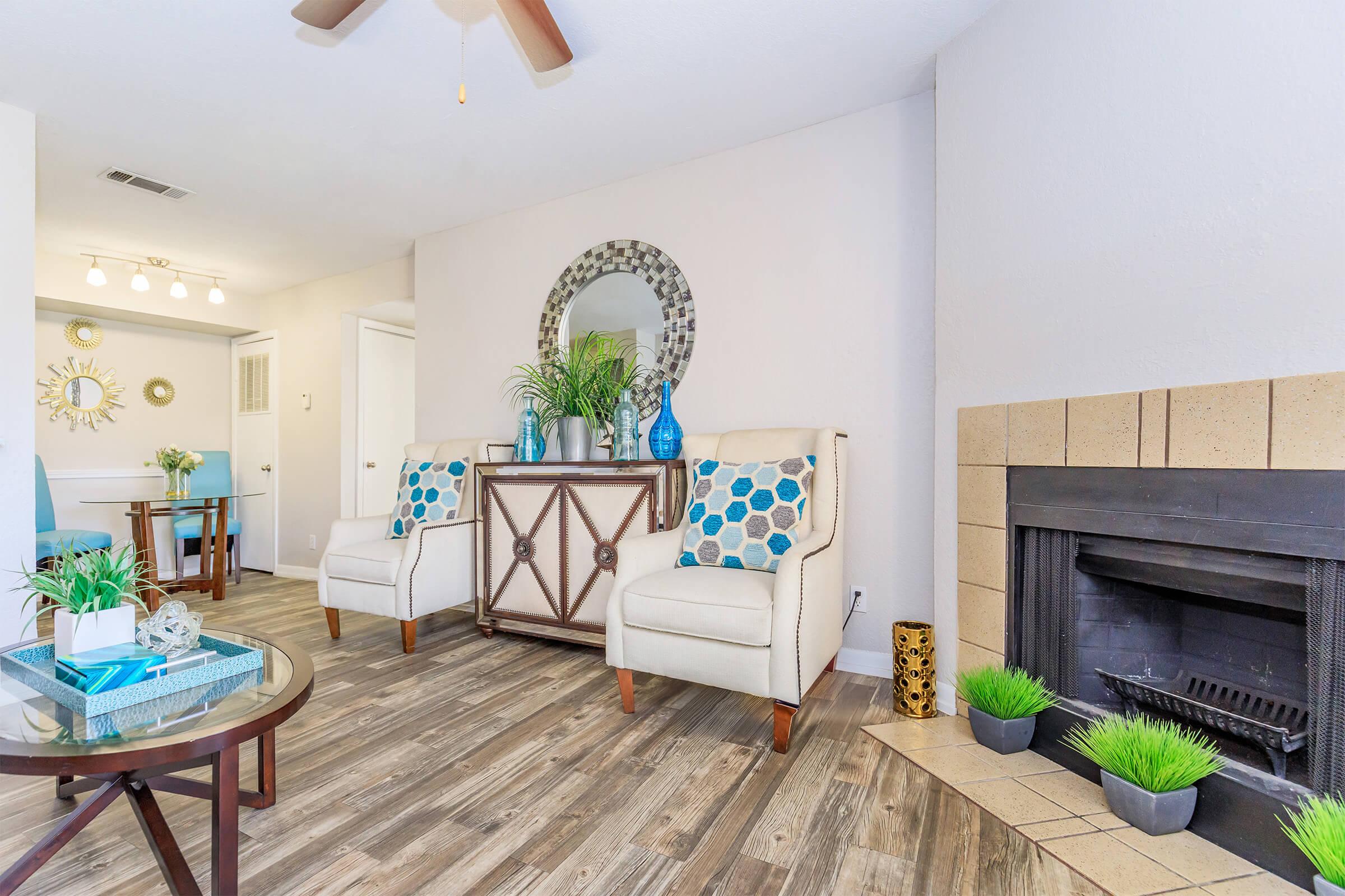 Bright and inviting living room featuring two elegant chairs with blue and white patterned pillows, a decorative wooden table in the center, and a cozy fireplace surrounded by light-colored tiles. Plants and decorative items enhance the warm atmosphere, with natural light illuminating the space.