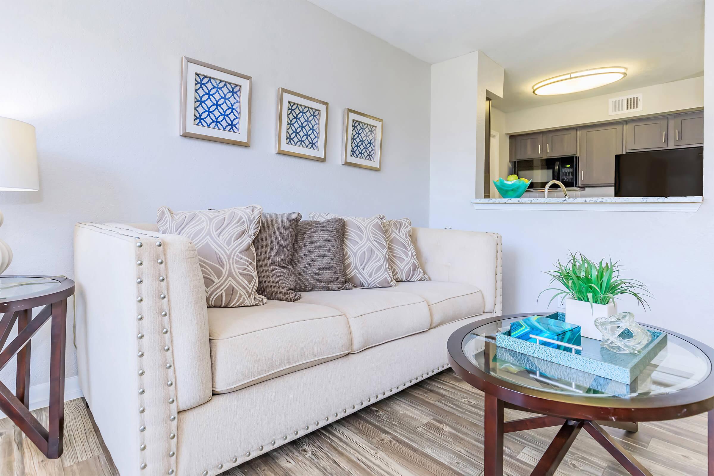 A cozy living room featuring a light-colored couch with decorative pillows, a round glass coffee table, and a small plant. The walls are adorned with framed artworks, and a kitchen area is visible in the background with modern appliances and a light fixture above.