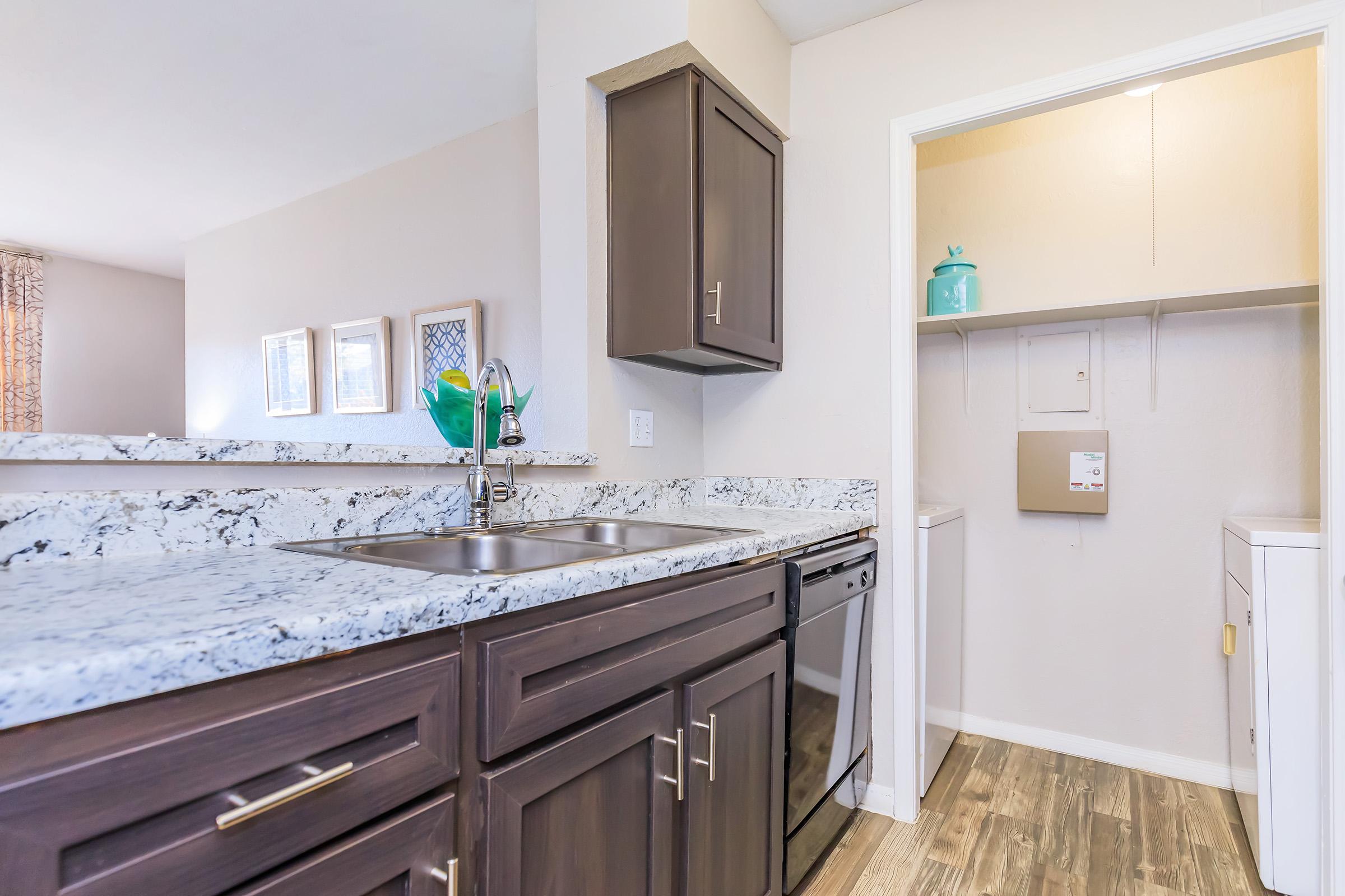 A modern kitchen featuring dark wood cabinets, a granite countertop, and a stainless steel sink. To the right, there's a laundry area with a washer and dryer. The walls are painted light, enhancing the bright, open atmosphere. Decorative frames are visible on the wall in the background.