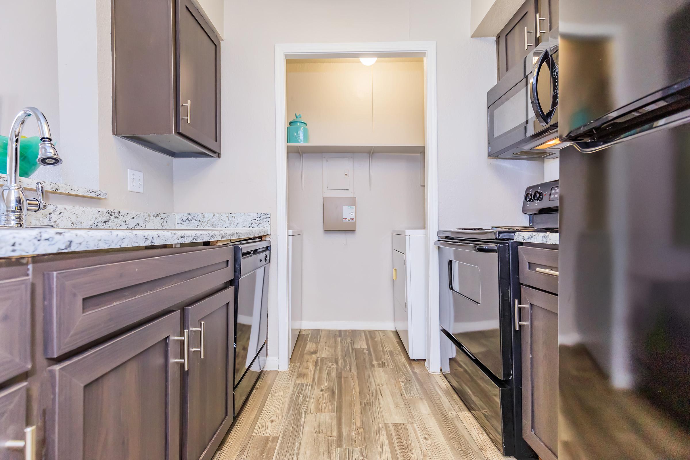 A spacious kitchen featuring dark cabinetry and granite countertops, with a view into a laundry nook. The kitchen is equipped with modern appliances, including a stove and dishwasher, and has a warm, inviting atmosphere highlighted by wood-look flooring.