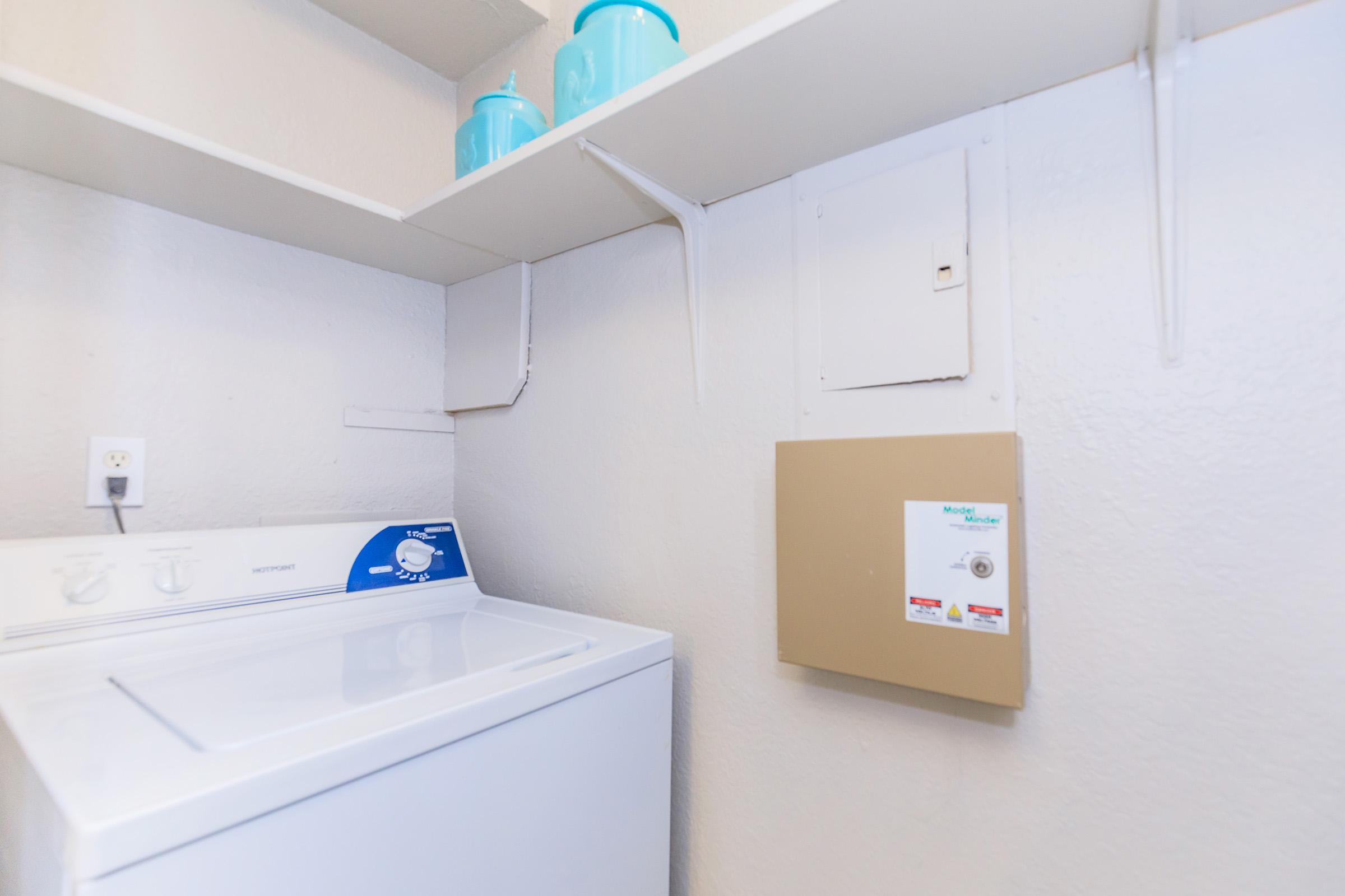 A clean laundry room featuring a white washing machine on the left, with shelves above holding three blue containers. The wall is painted light gray, and there is a beige electrical panel mounted on the wall, adjacent to the washing machine.