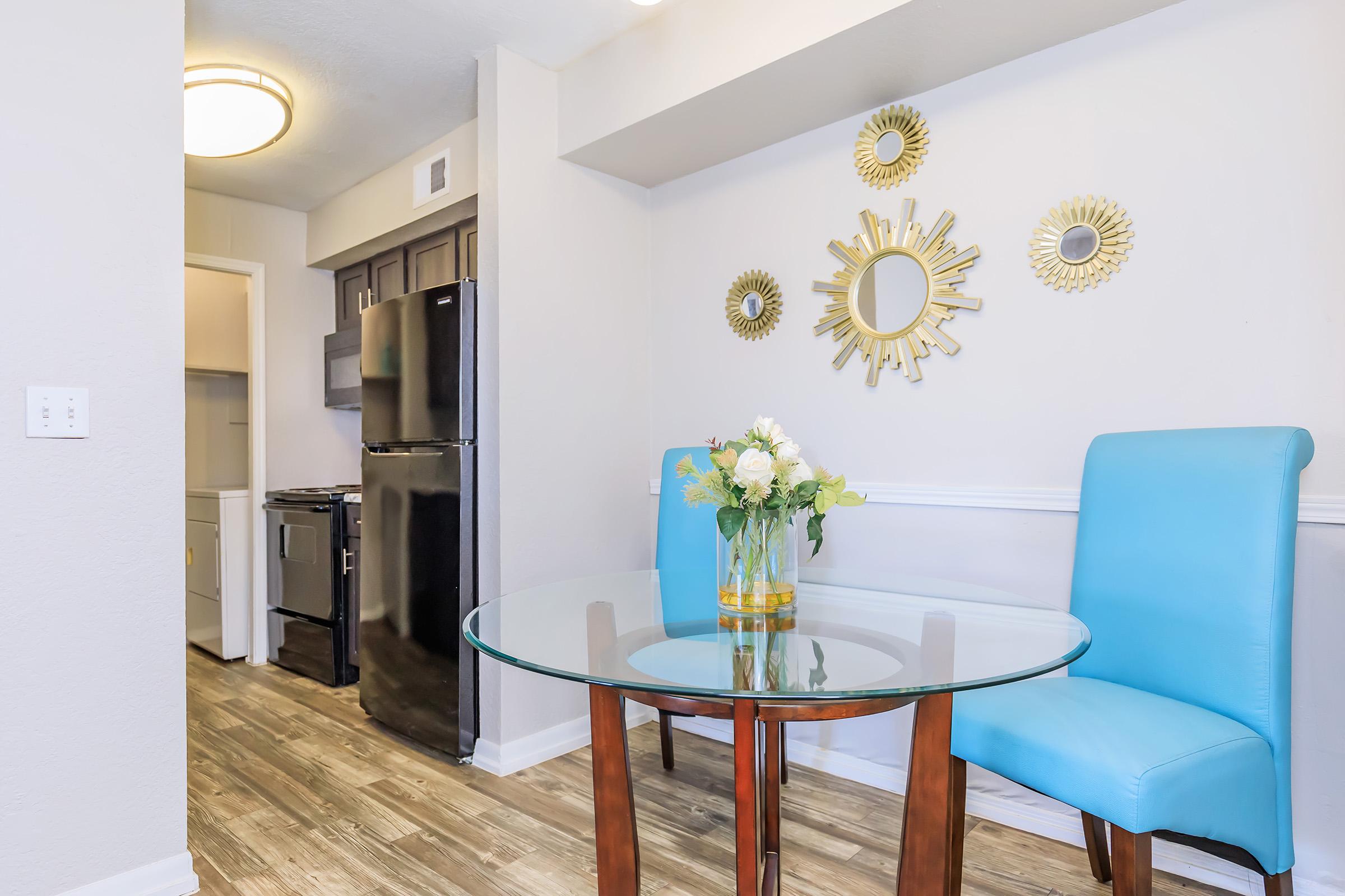 A modern dining area featuring a round glass table with a wooden base, surrounded by two light blue chairs. On the table is a vase of white flowers. The background shows a kitchen with a black refrigerator and cabinets, and decorative sunburst mirrors on the wall.