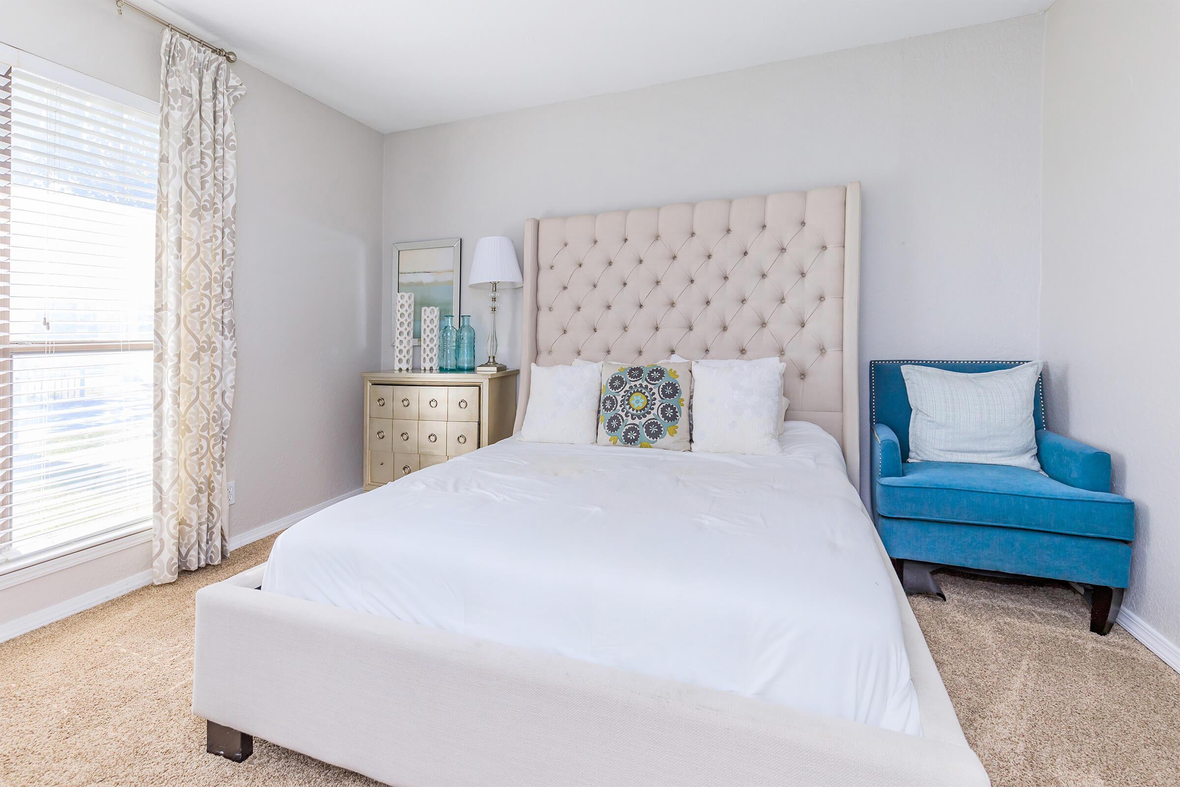 A bright, modern bedroom featuring a tufted beige bed with white linens, a decorative pillow, and a matching headboard. There’s a stylish light blue armchair in the corner, a mirrored nightstand with decorative glass items, and large windows with sheer curtains allowing natural light to fill the space.