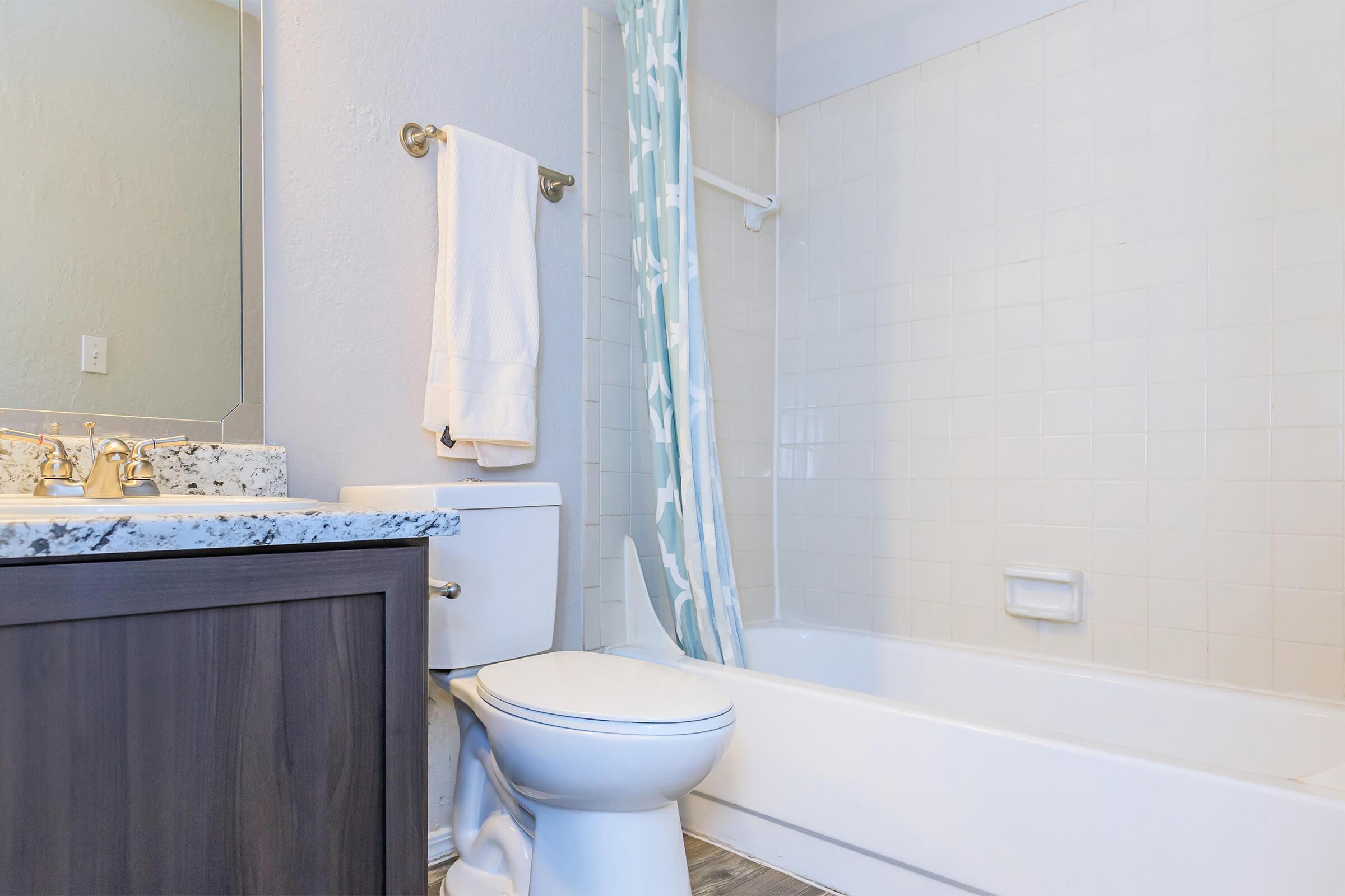 A clean and modern bathroom featuring a white bathtub, toilet, and a granite countertop sink. The shower area has a light blue patterned curtain, and there are white towels hanging on a towel rack. The walls are painted light grey, creating a bright and spacious atmosphere.