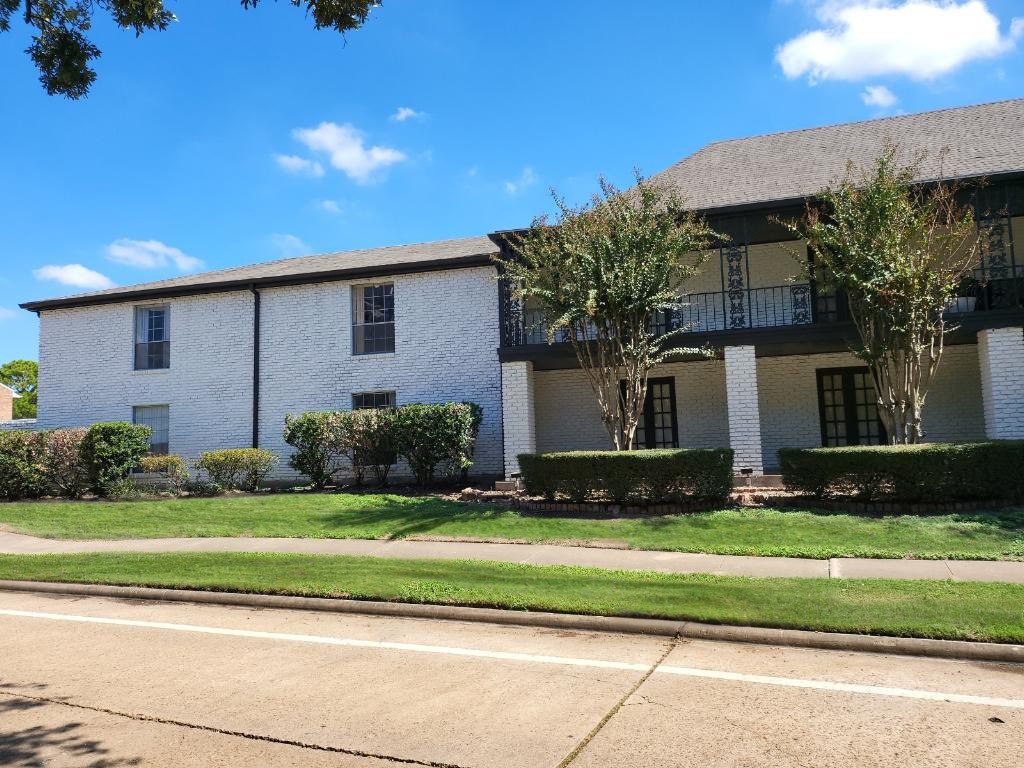 A two-story white brick apartment building with black railings, surrounded by lush greenery and neatly trimmed hedges. The blue sky in the background features a few fluffy clouds, and a sidewalk runs along the front of the property.