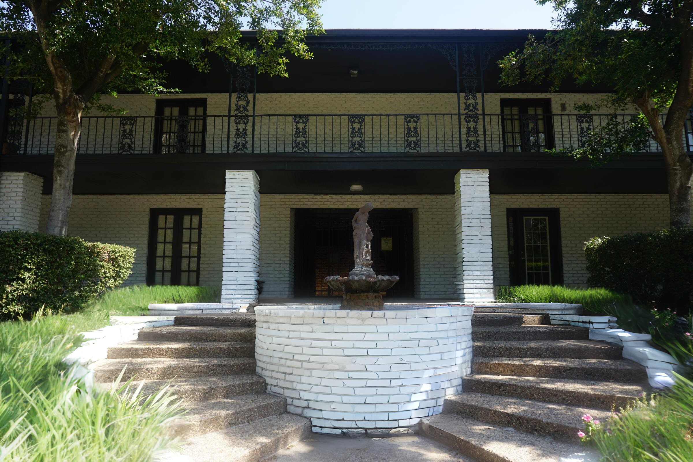 A stately two-story building with a dark exterior and decorative wrought iron railings. In front, a circular stone fountain is surrounded by landscaped greenery and stone steps leading up to the entrance. Two well-manicured trees flank either side of the steps, enhancing the elegant setting.