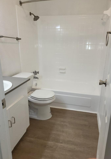 A clean, modern bathroom featuring a white tub and shower with tiled walls, a toilet, and a small sink with a gray countertop. The floor has a wood-like appearance, and there are white walls with minimal fixtures. A door leads to the bathroom, adding to the uncluttered look.