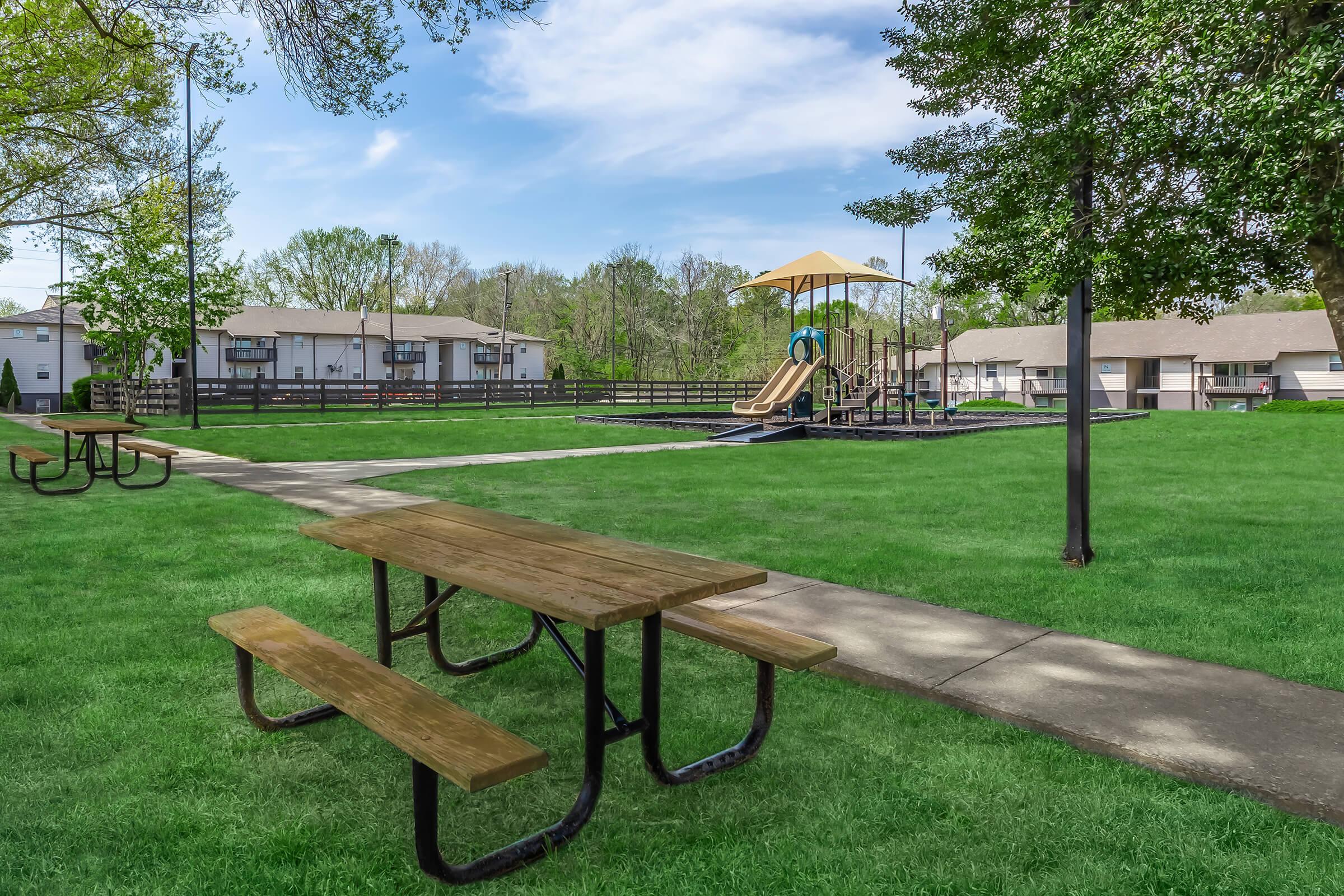 A spacious park featuring green grass, a picnic table in the foreground, and a playground in the background with a slide and climbing structure. Nearby, there are several residential buildings and trees, creating a serene outdoor environment.