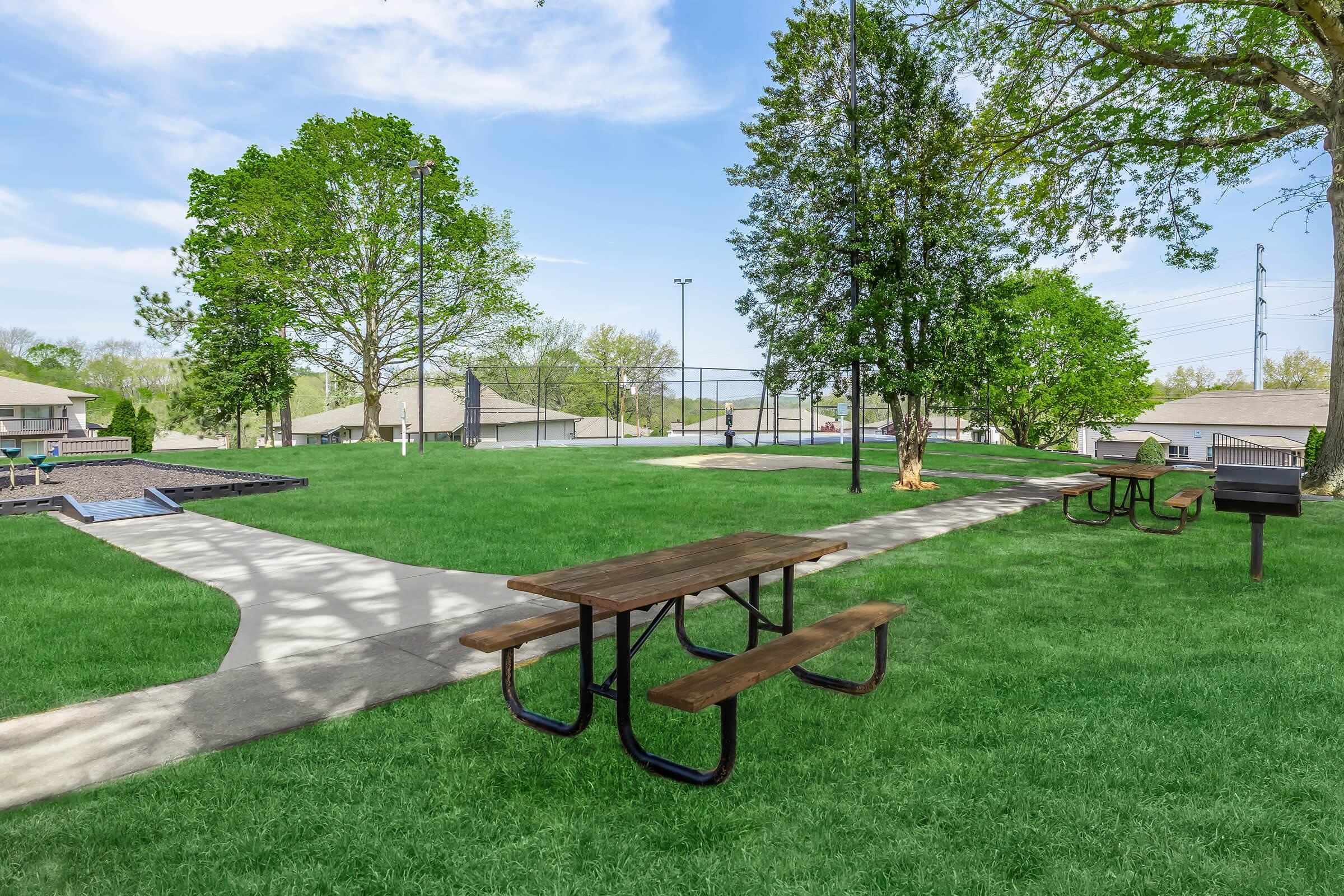A peaceful park scene featuring a grassy area with several brown picnic tables, a walking path, and a grill. Lush trees line the background, and a sports court is visible in the distance under a clear blue sky. Ideal for outdoor gatherings and leisure activities.