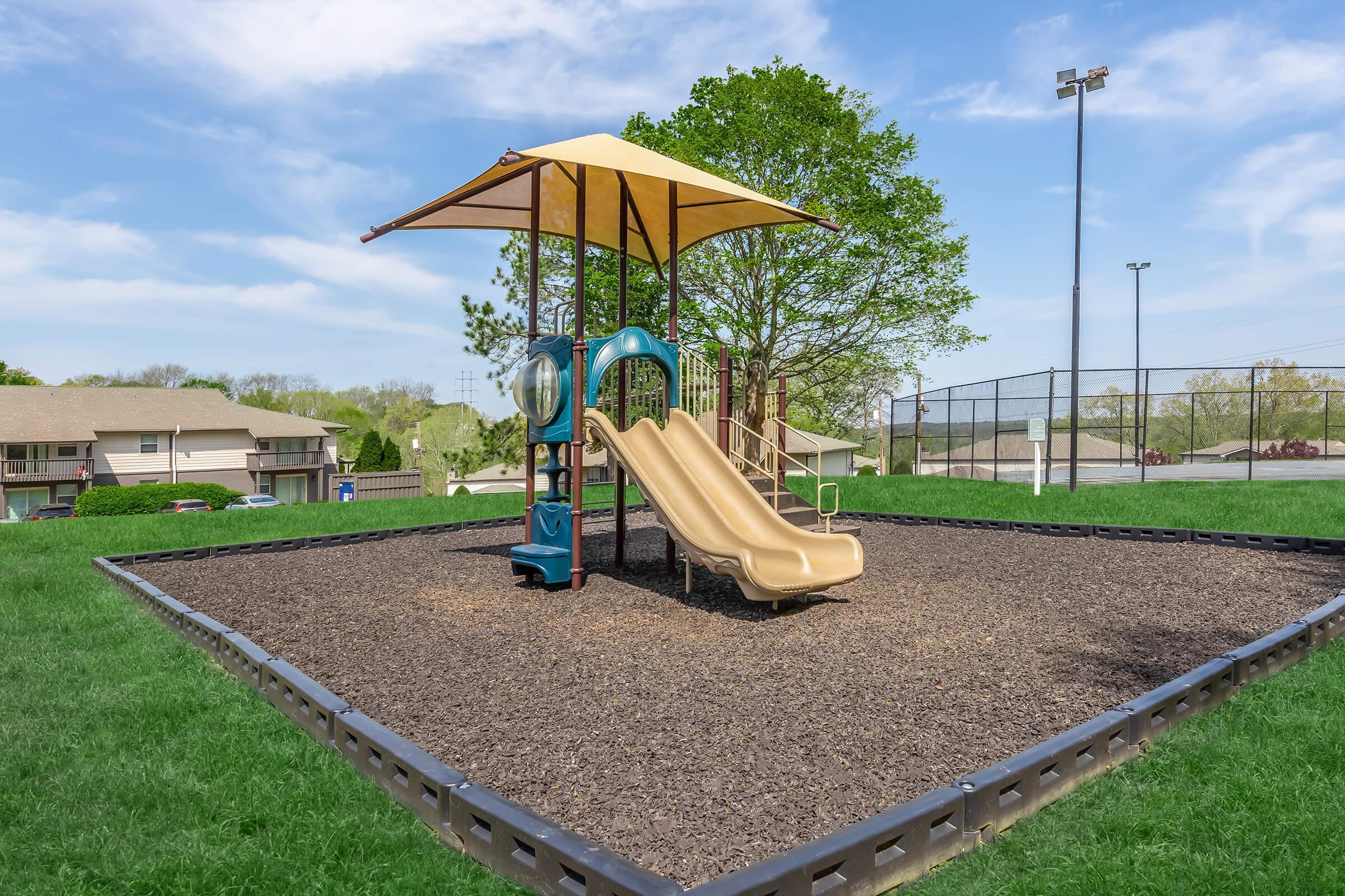 A playground featuring a yellow and brown slide under a shaded structure, surrounded by green grass and bordered by a rubberized black perimeter. In the background, there are residential buildings and a fenced basketball court under a clear blue sky.