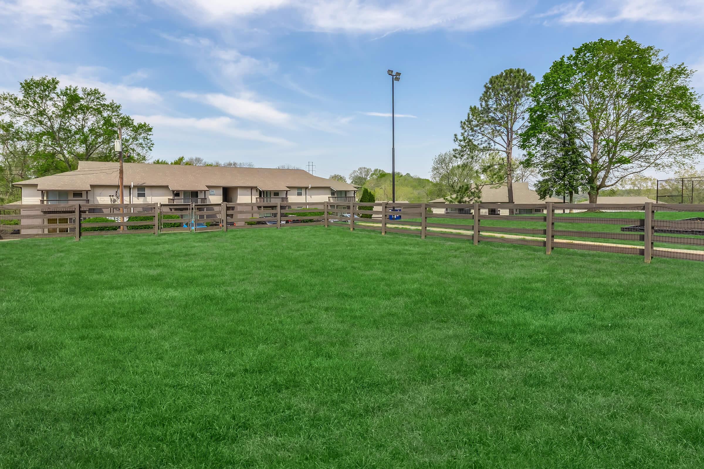 A spacious green lawn enclosed by wooden fencing, with several trees and a residential building in the background. The sky is clear with a few clouds, and there’s a light pole visible. The scene suggests a peaceful outdoor area, possibly a park or community space.