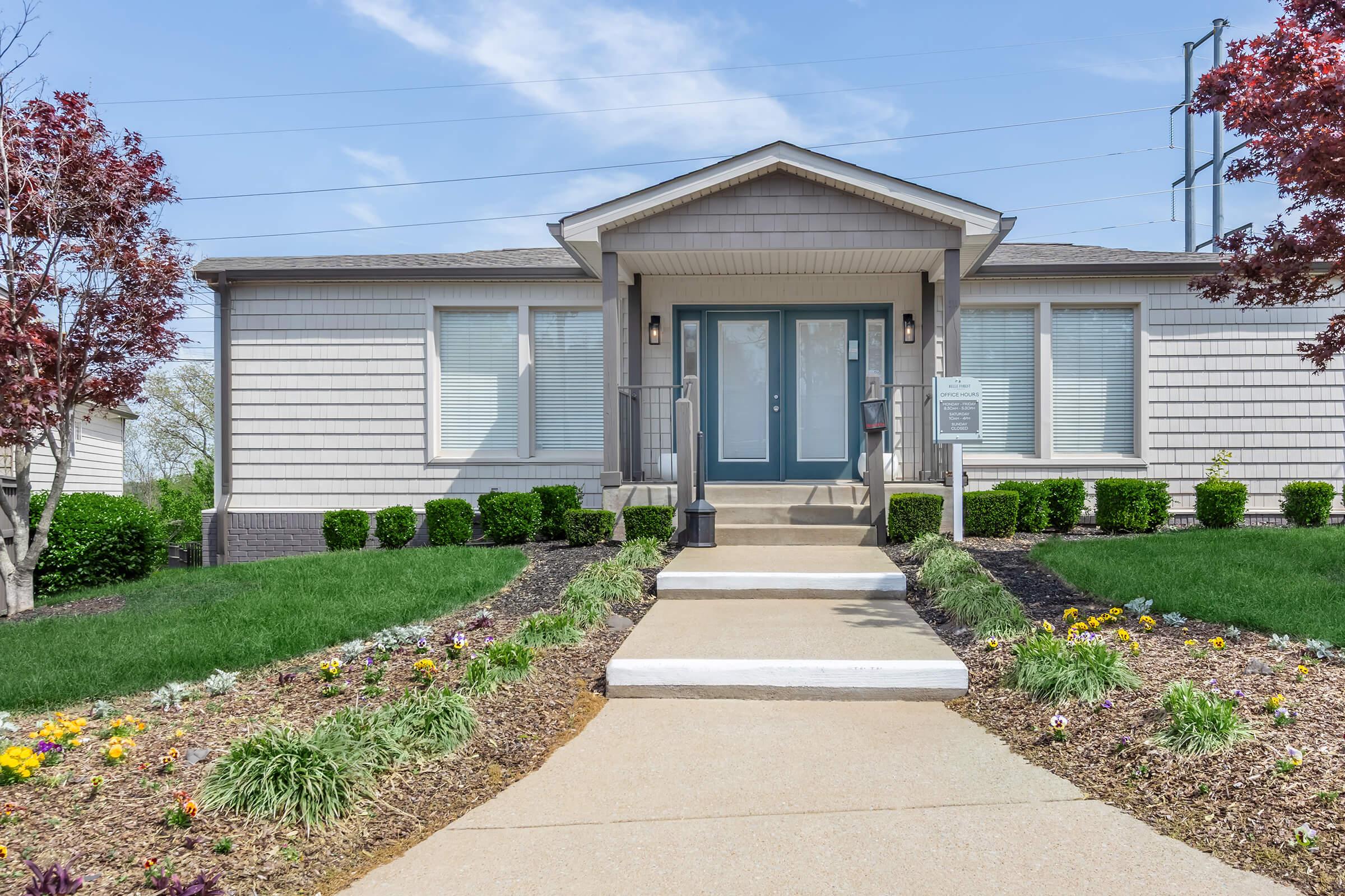 A modern single-story house with a light gray exterior and a welcoming front entrance. The pathway is lined with manicured grass and colorful flower beds. There are symmetrical shrubs on either side of the entrance, and the sky is clear with a few clouds.