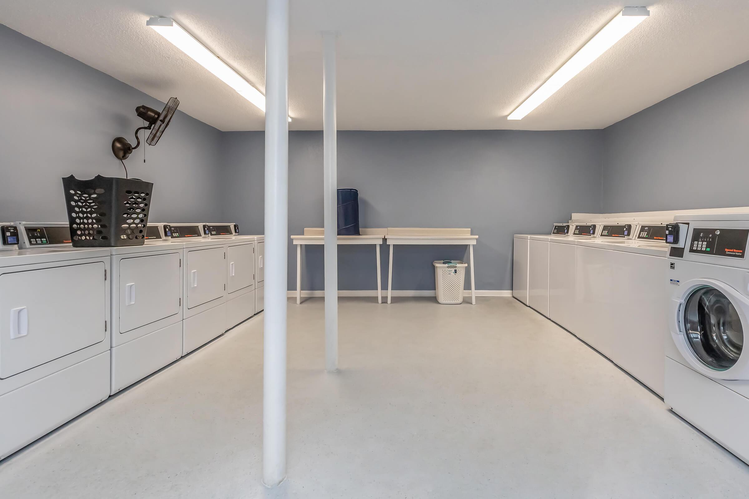 A clean and modern laundry room featuring multiple white washing machines and dryers arranged along the walls. The space is well-lit with overhead lights, has a blue wall, and includes a fold-out table, a laundry basket, and a wall-mounted item organizer. The floor is light-colored and the area is tidy.