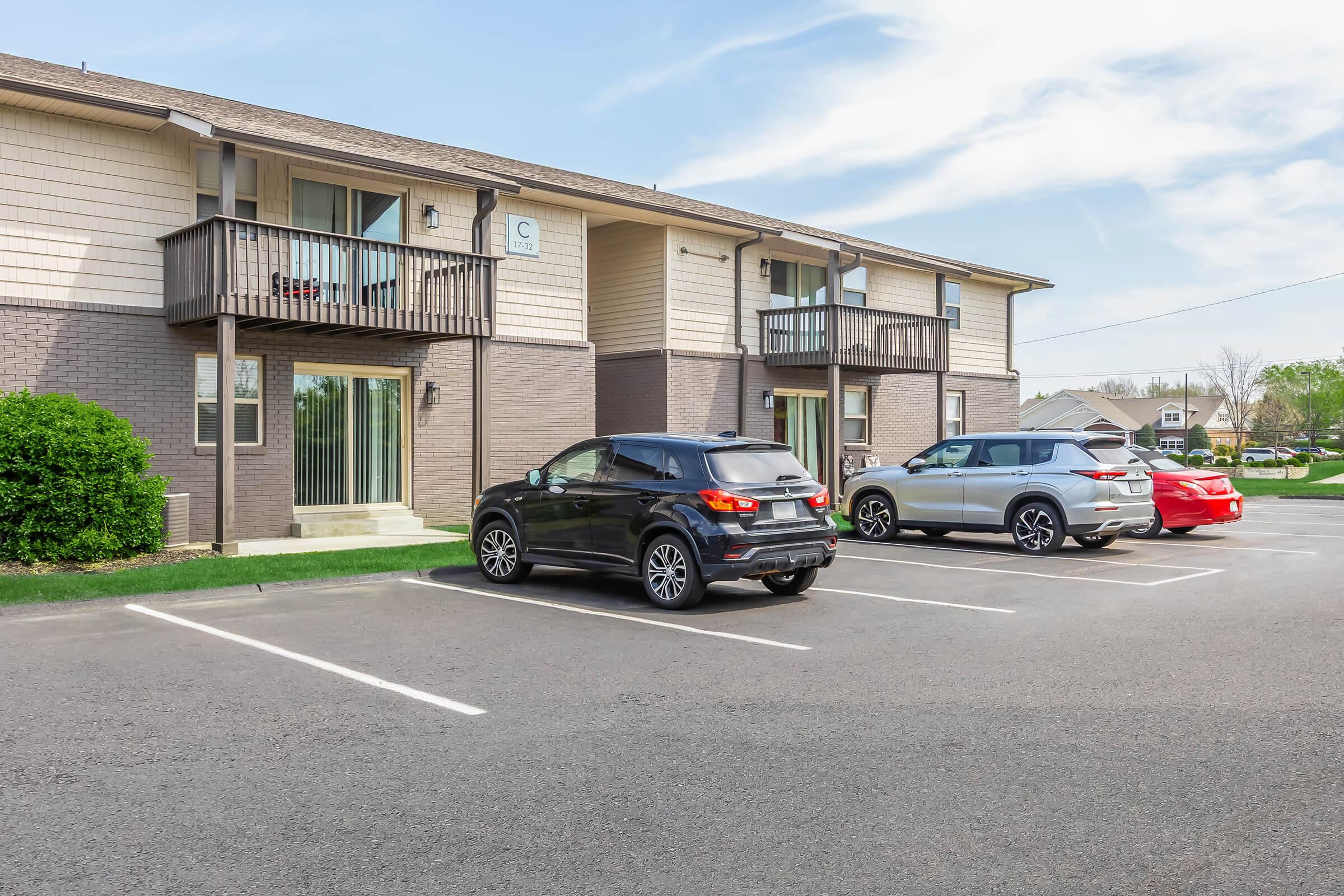 Two apartment buildings with a parking lot in the foreground. A black SUV and a silver car are parked in front of the buildings, along with a red car further down the lot. Green grass and trees are visible in the background under a clear blue sky.
