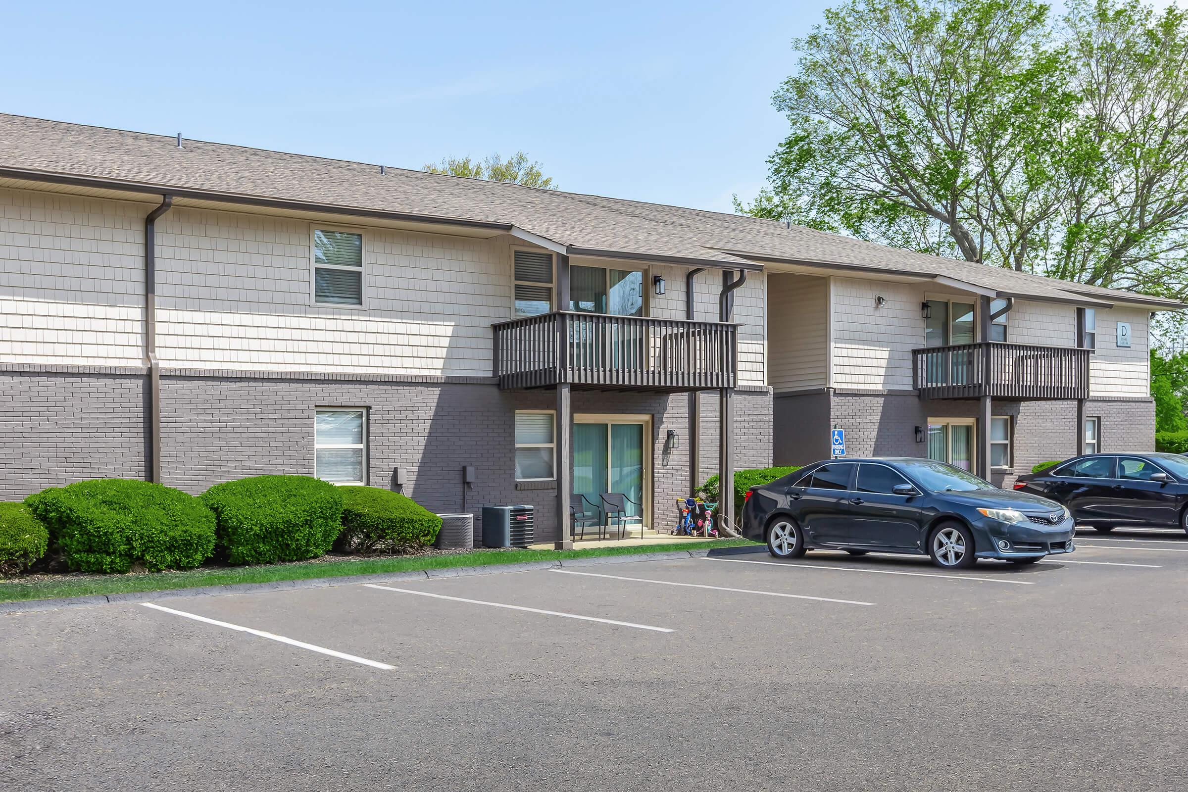 A two-story apartment building with a brick and siding exterior, featuring balconies. A black car is parked in the foreground, with a row of parking spaces visible. Green shrubs line the building, and trees are present in the background under a clear blue sky.