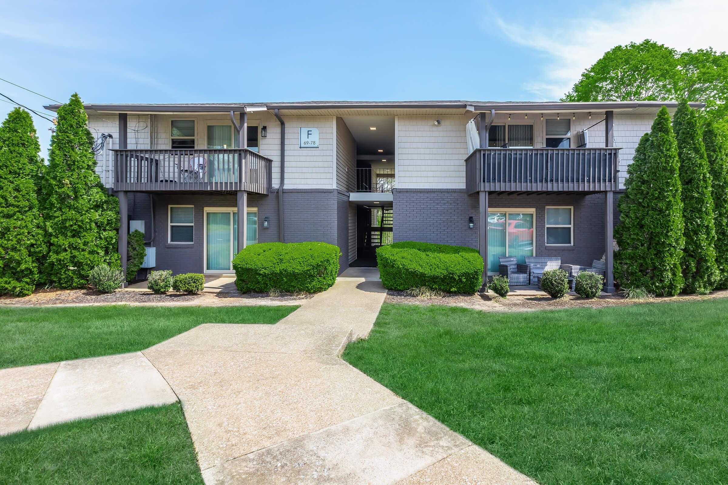 Two-story apartment building with a gray exterior, featuring multiple entrances with balconies. Lush green bushes and neatly trimmed grass surround the building. A concrete walkway leads to the entrances, set against a clear blue sky.