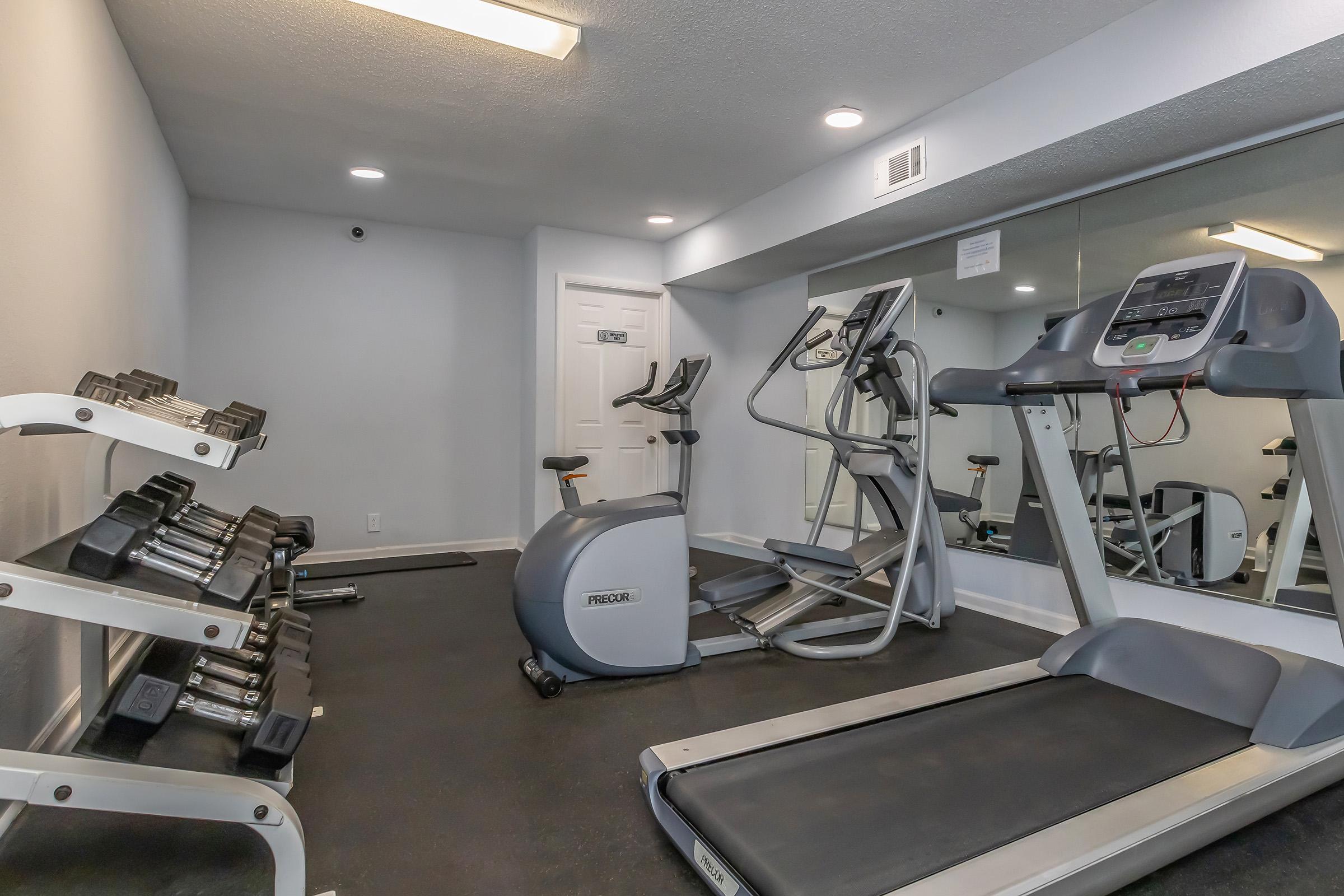 A small gym area featuring an elliptical machine, a treadmill, and a set of free weights on a rack. The space has light-colored walls, a mirror, and is well-lit, creating an inviting atmosphere for workouts.