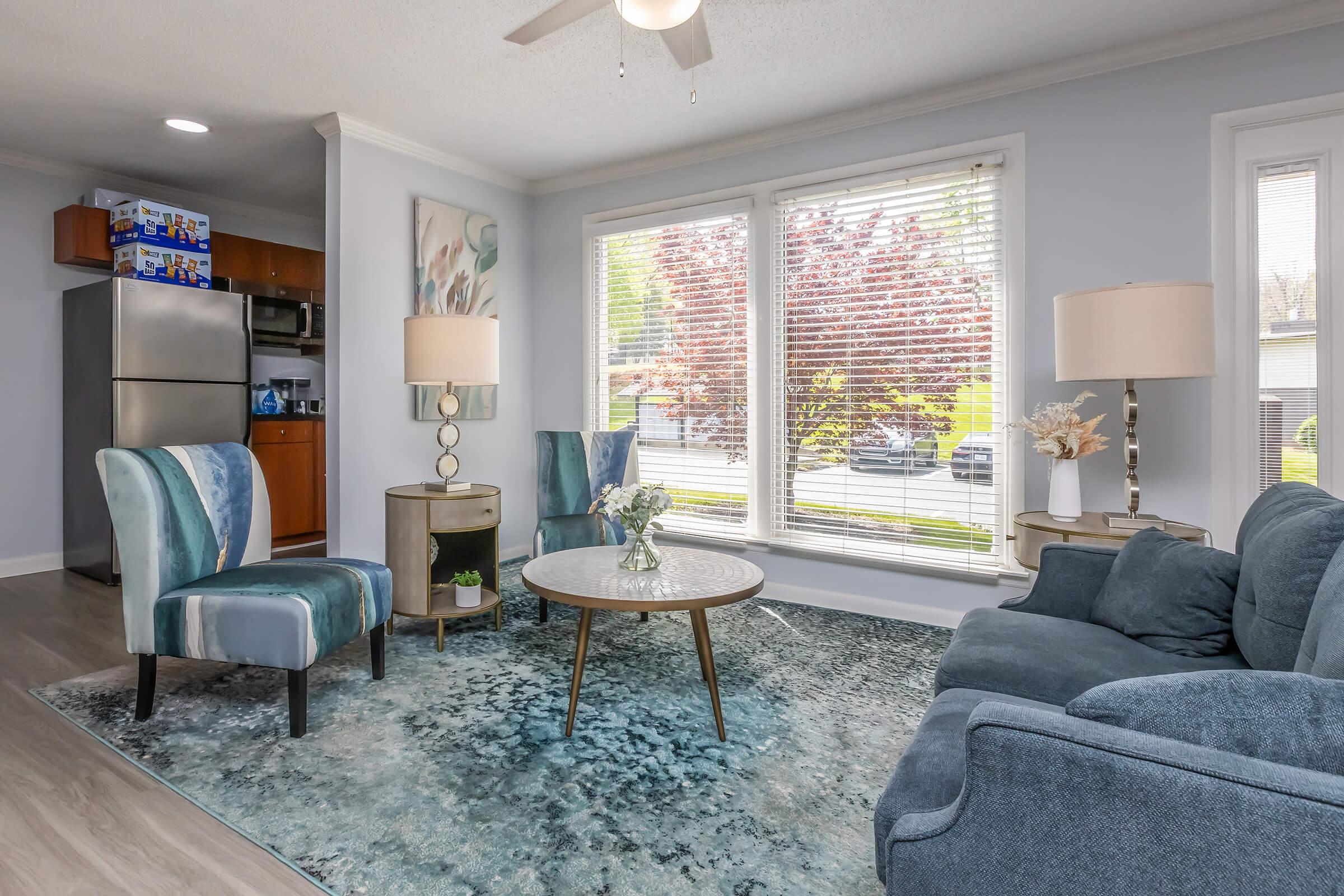 A cozy living room featuring a blue area rug, a round coffee table, a sofa, and two accent chairs. Large windows let in natural light and offer a view of a tree-lined street. Decor includes lamps and a floral arrangement on the table, creating a welcoming atmosphere.
