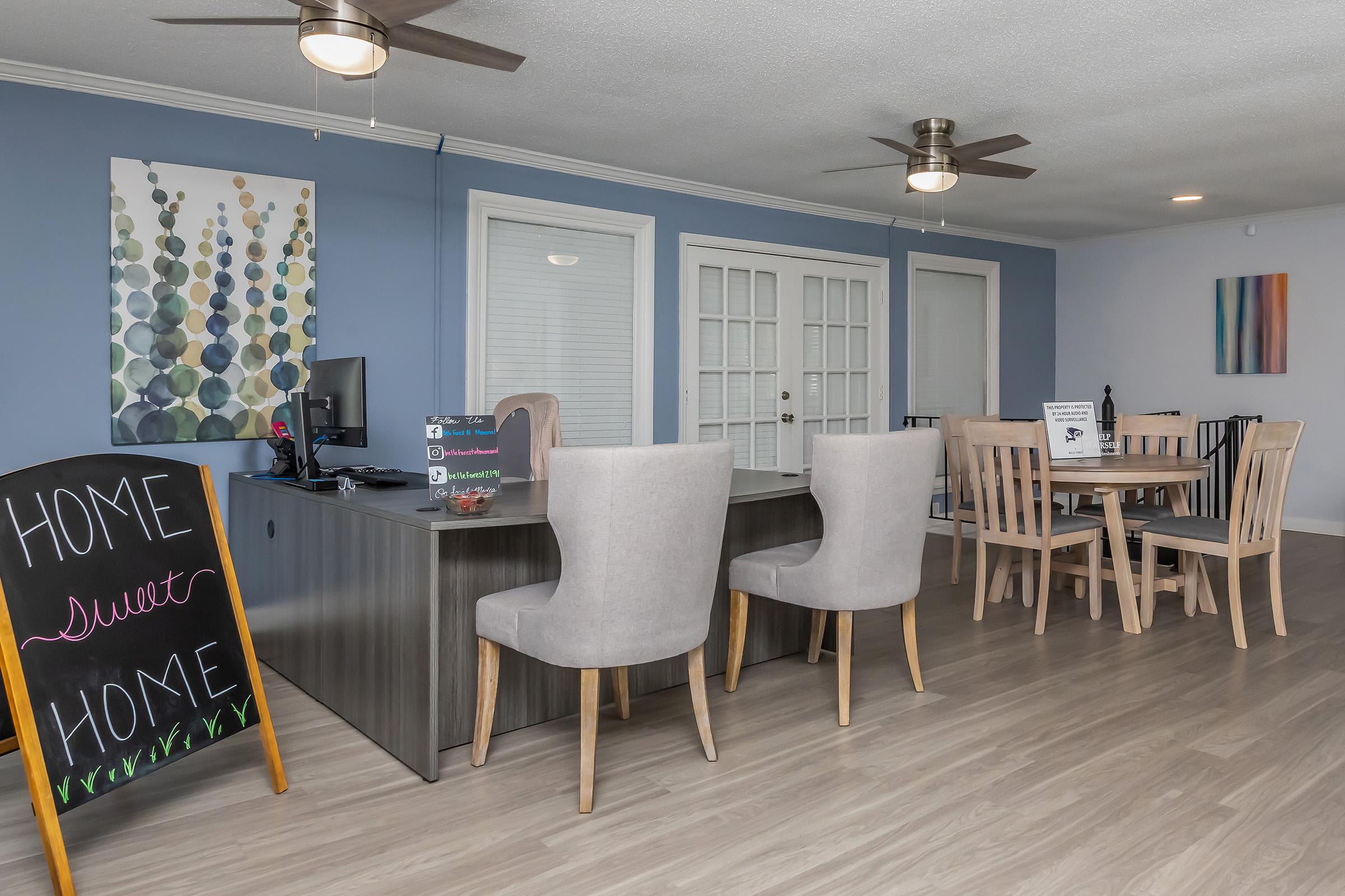 Modern interior of a welcoming reception area featuring a stylish desk with computer, two comfortable light-colored chairs, a dining table with wooden chairs, and decorative wall art. A chalkboard sign with "Home Sweet Home" is prominently displayed, adding a warm touch to the space.
