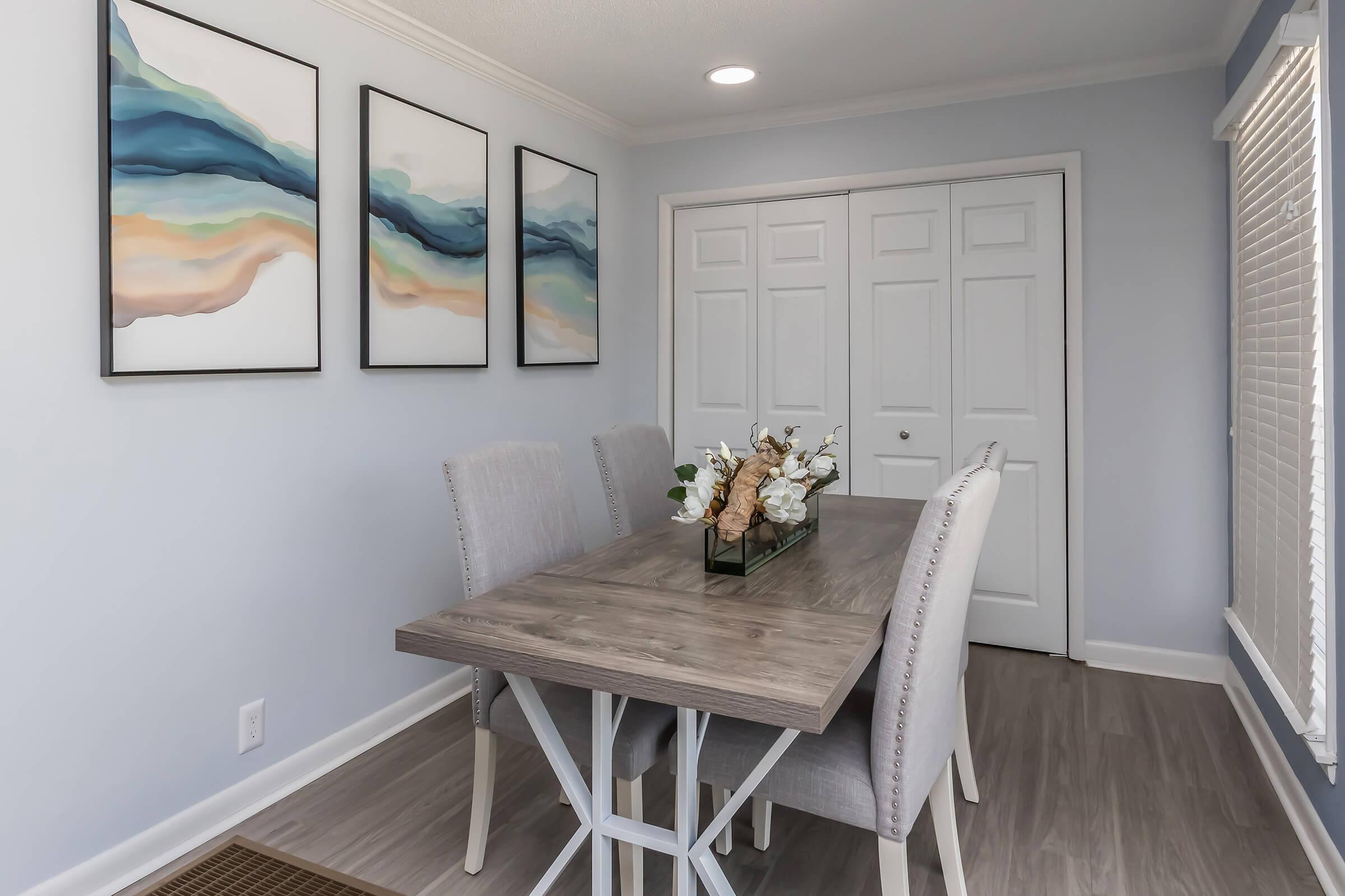 A modern dining area featuring a wooden table with four gray upholstered chairs. Above the table, there are three abstract paintings in soft colors. A glass vase with decorative flowers sits at the center of the table. The walls are painted light blue, and there is a closet with double doors.