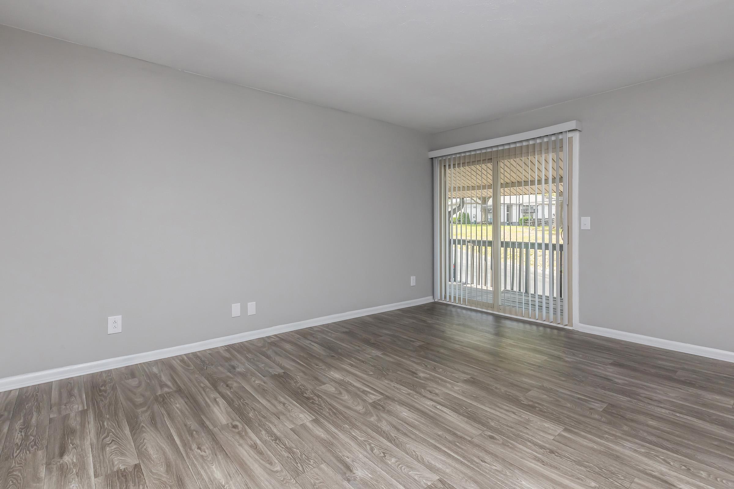 Empty room with light gray walls and wood-like flooring. A sliding glass door with vertical blinds leads to an outdoor area. There are two electrical outlets and no furniture present, creating an open and spacious feel. Natural light is visible through the door.
