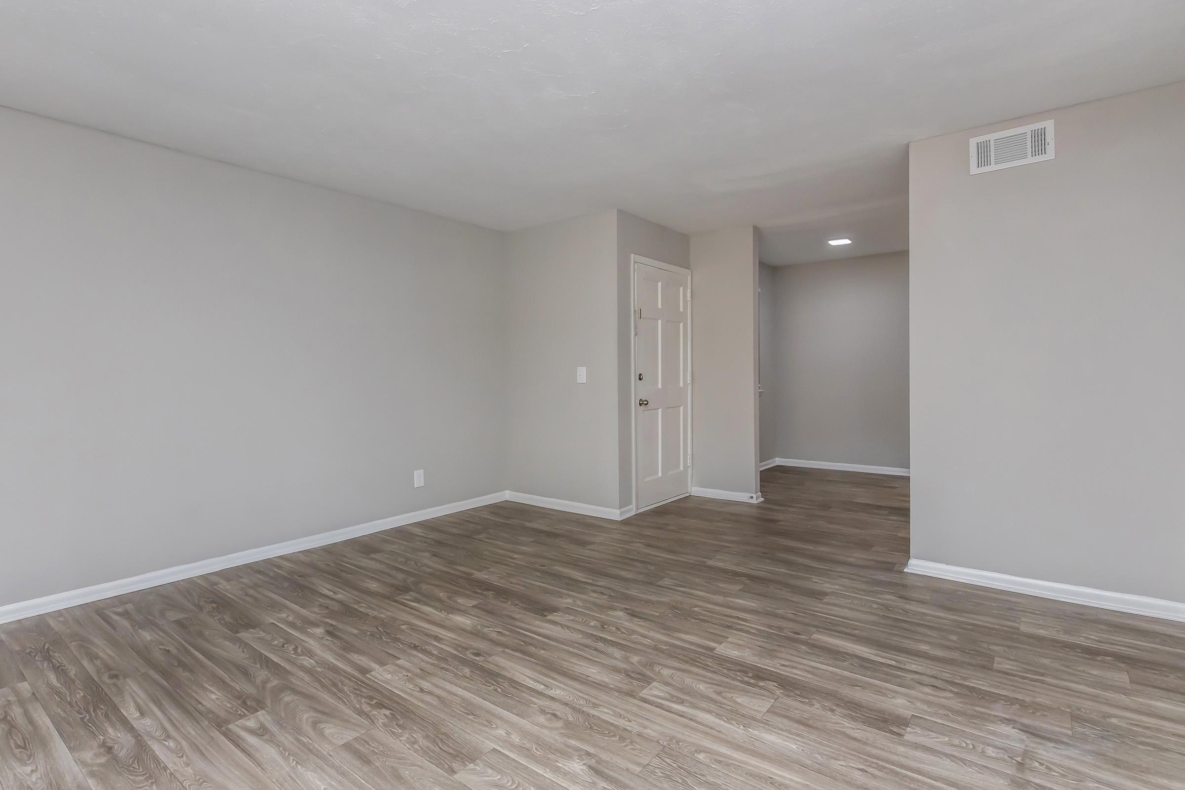 A spacious, empty living area with light gray walls and modern wood-style flooring. There is a doorway visible, leading to another room, and the overall lighting is bright and neutral, creating an airy atmosphere.