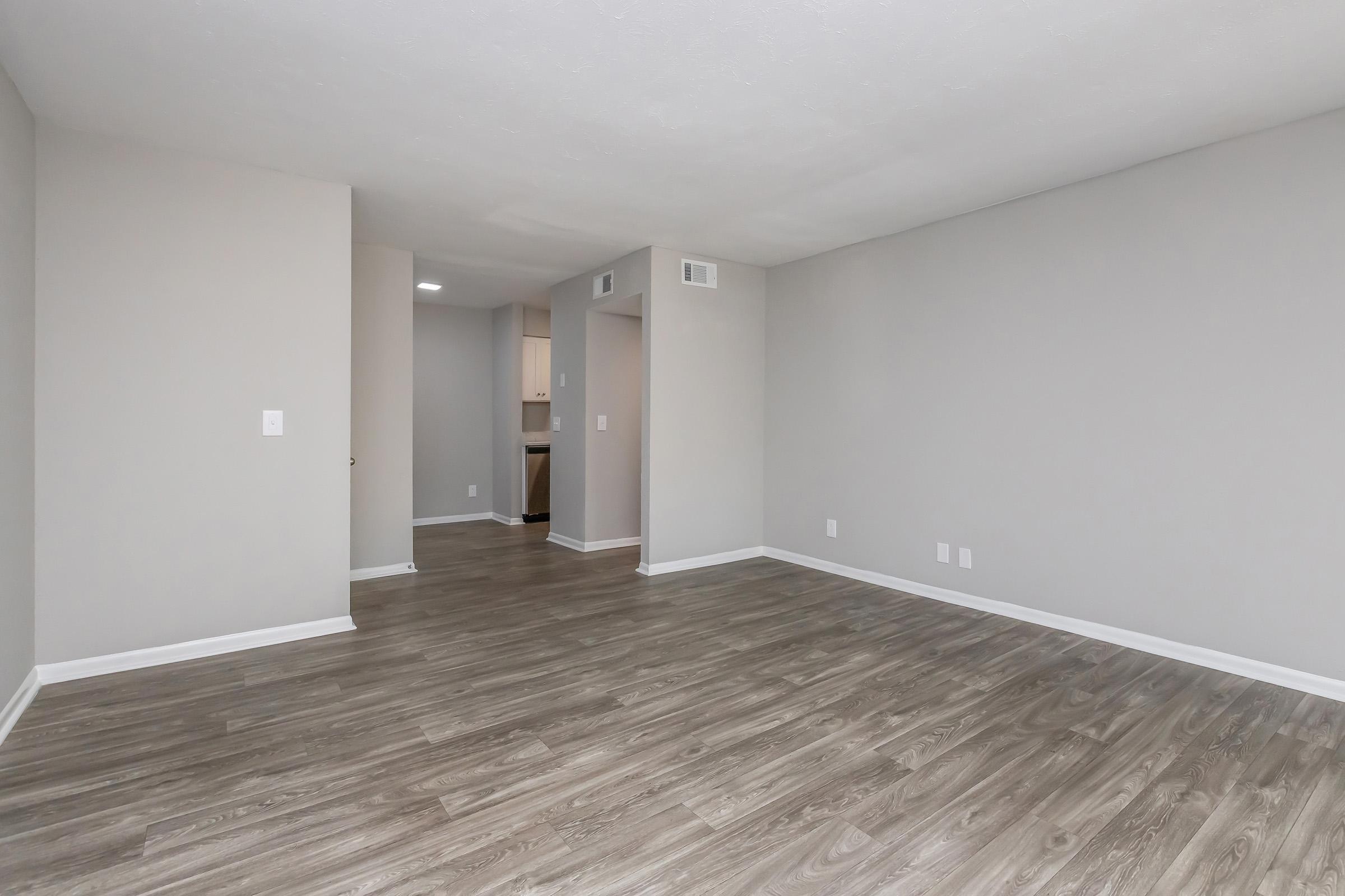 An empty room featuring light gray walls and a wide expanse of laminate flooring. The space includes a doorway leading to a kitchen area in the background, with no furniture present, creating an open and airy atmosphere. Natural light enhances the simplicity of the interior design.