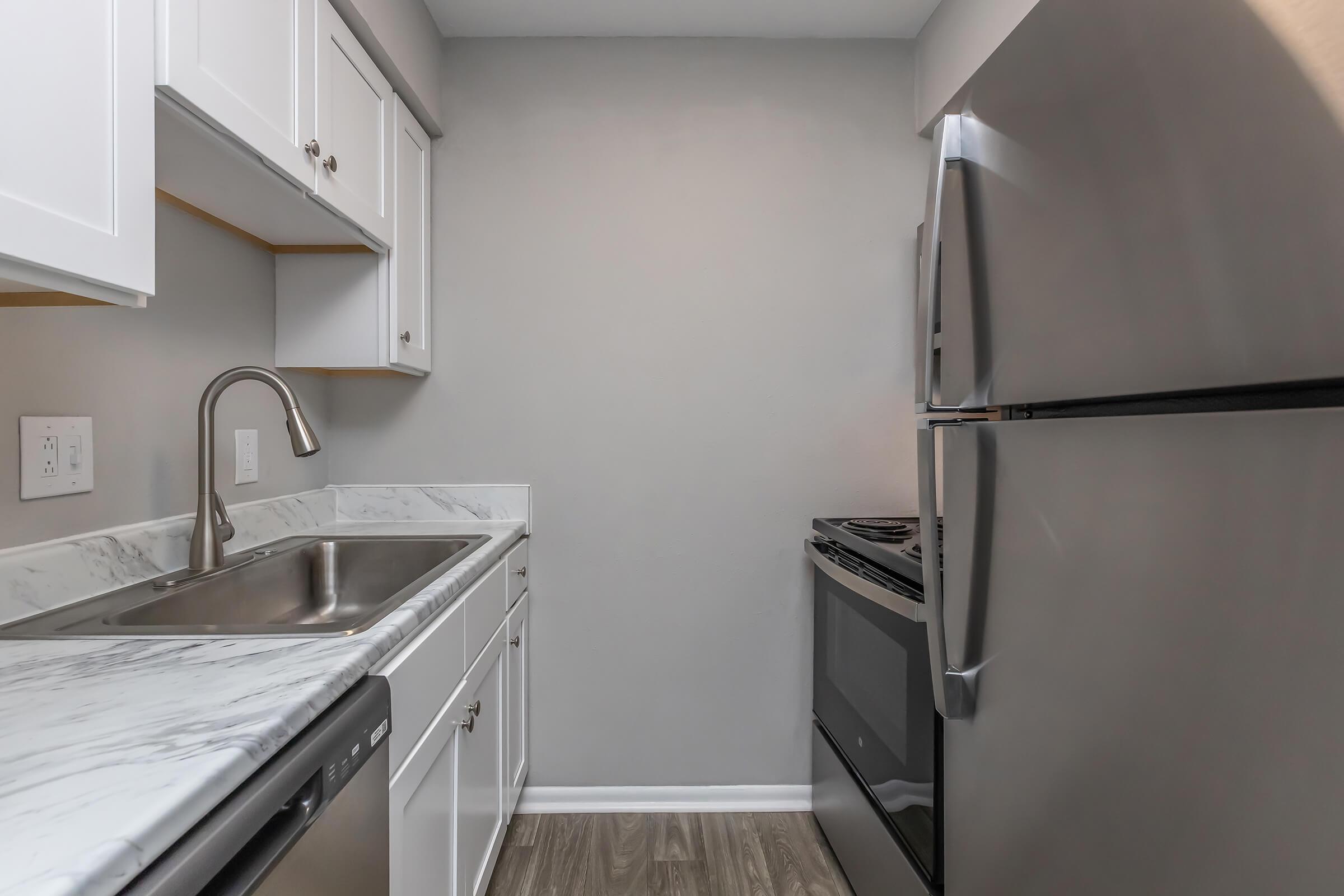 A modern kitchen featuring white cabinets, a stainless steel refrigerator, a stainless steel sink with a single handle faucet, and a black electric stove. The countertop is marble, and the flooring is wood-like. The walls are painted in a soft gray, creating a contemporary and clean aesthetic.