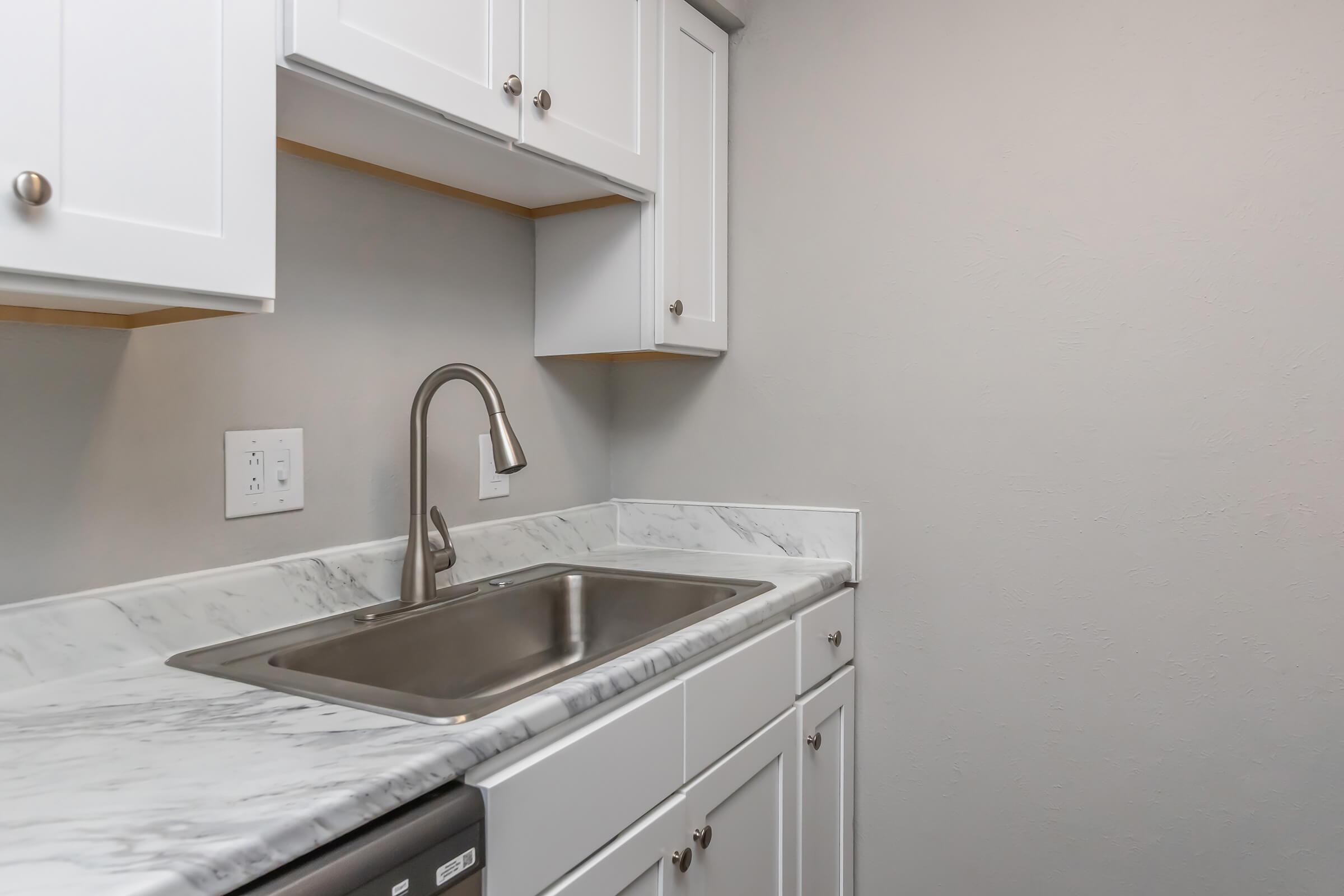 A modern kitchen with white cabinets, a gray countertop, and a stainless steel sink. The walls are painted light gray, creating a clean and minimalist look. There is also a dishwasher visible next to the sink.