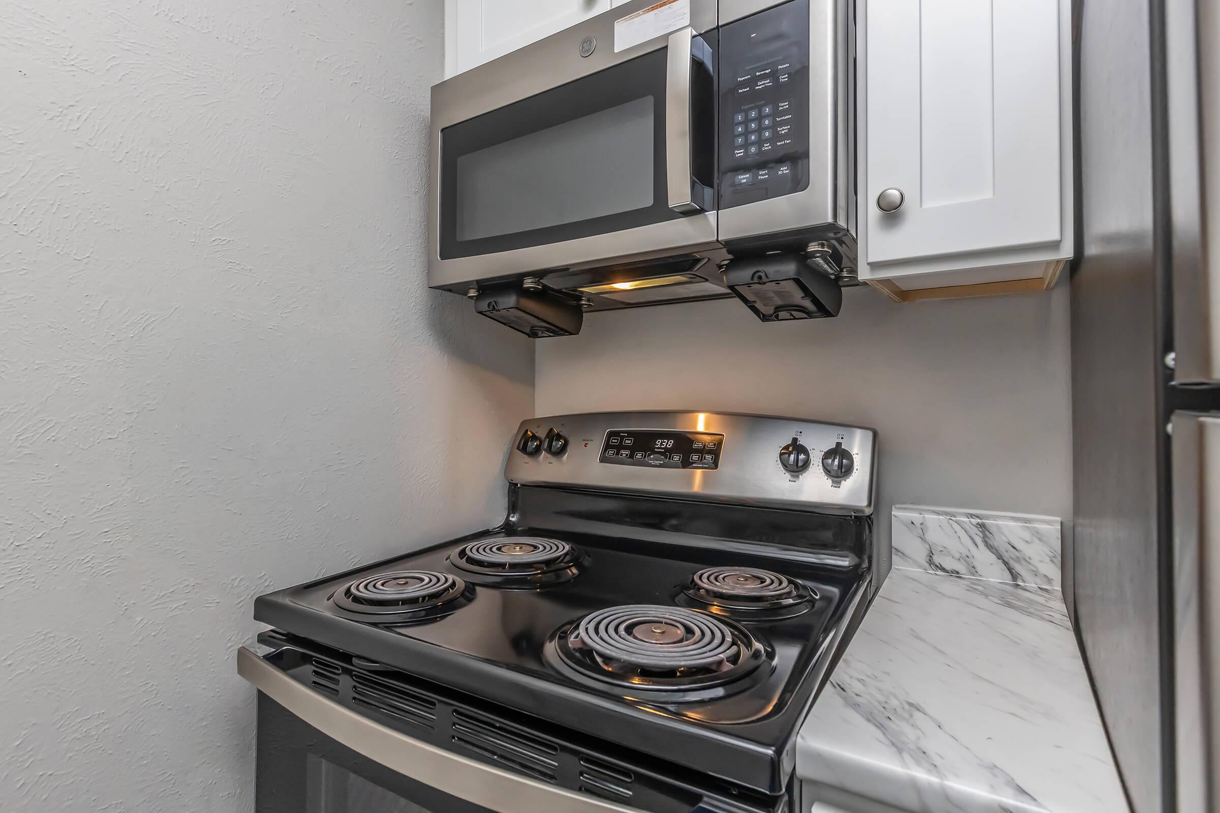 A modern kitchen featuring a black stove with four burners, a stainless steel microwave mounted above it, and white cabinets. The stove has control knobs and a digital display. A marble countertop is visible beneath the stove, complemented by a gray wall background.