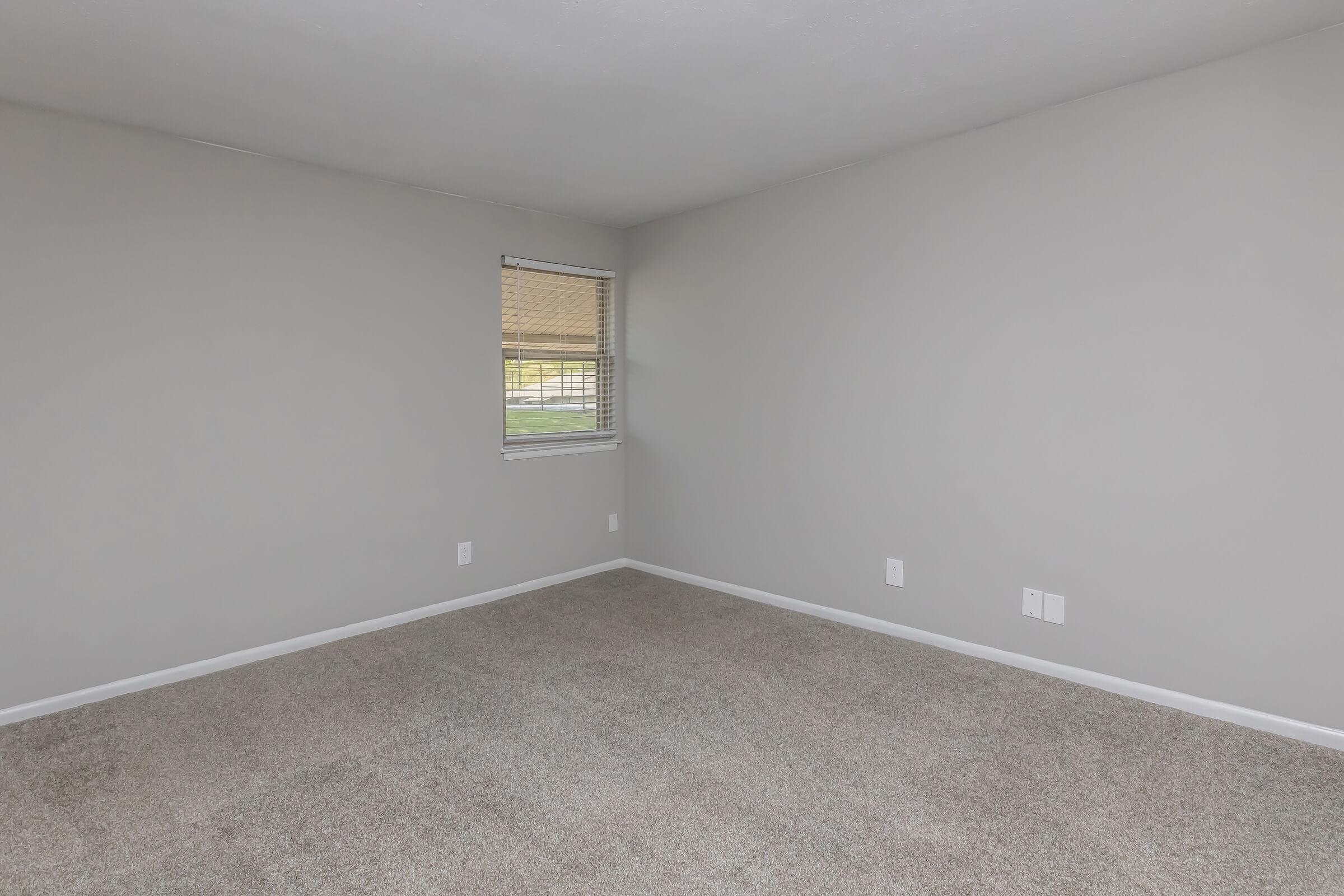 A vacant room with light gray walls and carpet. A window with blinds allows natural light, and there are two electrical outlets visible on one wall. The space is clean and unfurnished, creating a minimalist appearance.