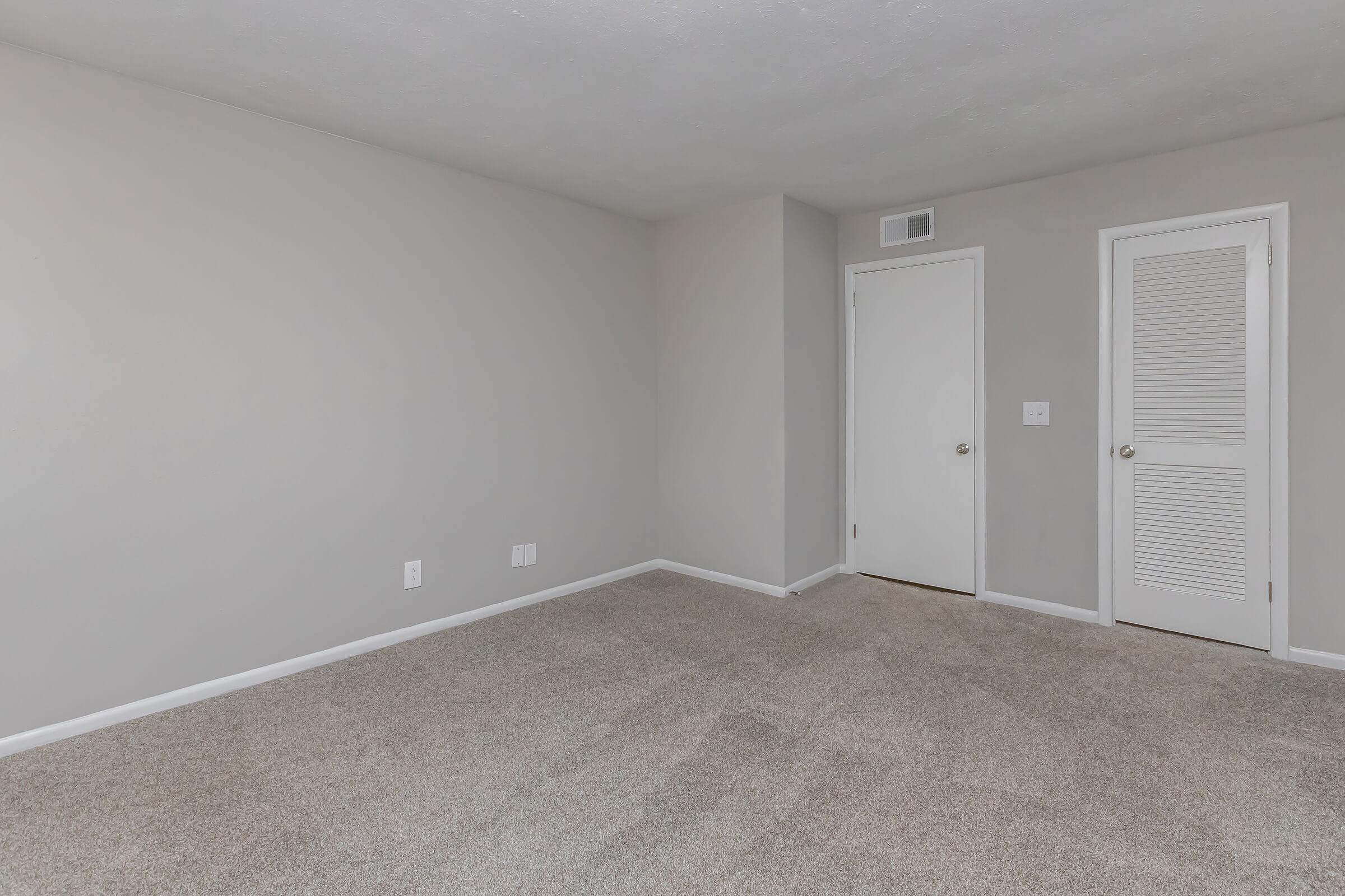 Empty room with light gray walls and carpeted floor. One door is visible on the right, leading to a closet with louvers. The other door is plain and leads to another space. The room has no furniture or decorations, creating a spacious and minimalist atmosphere.