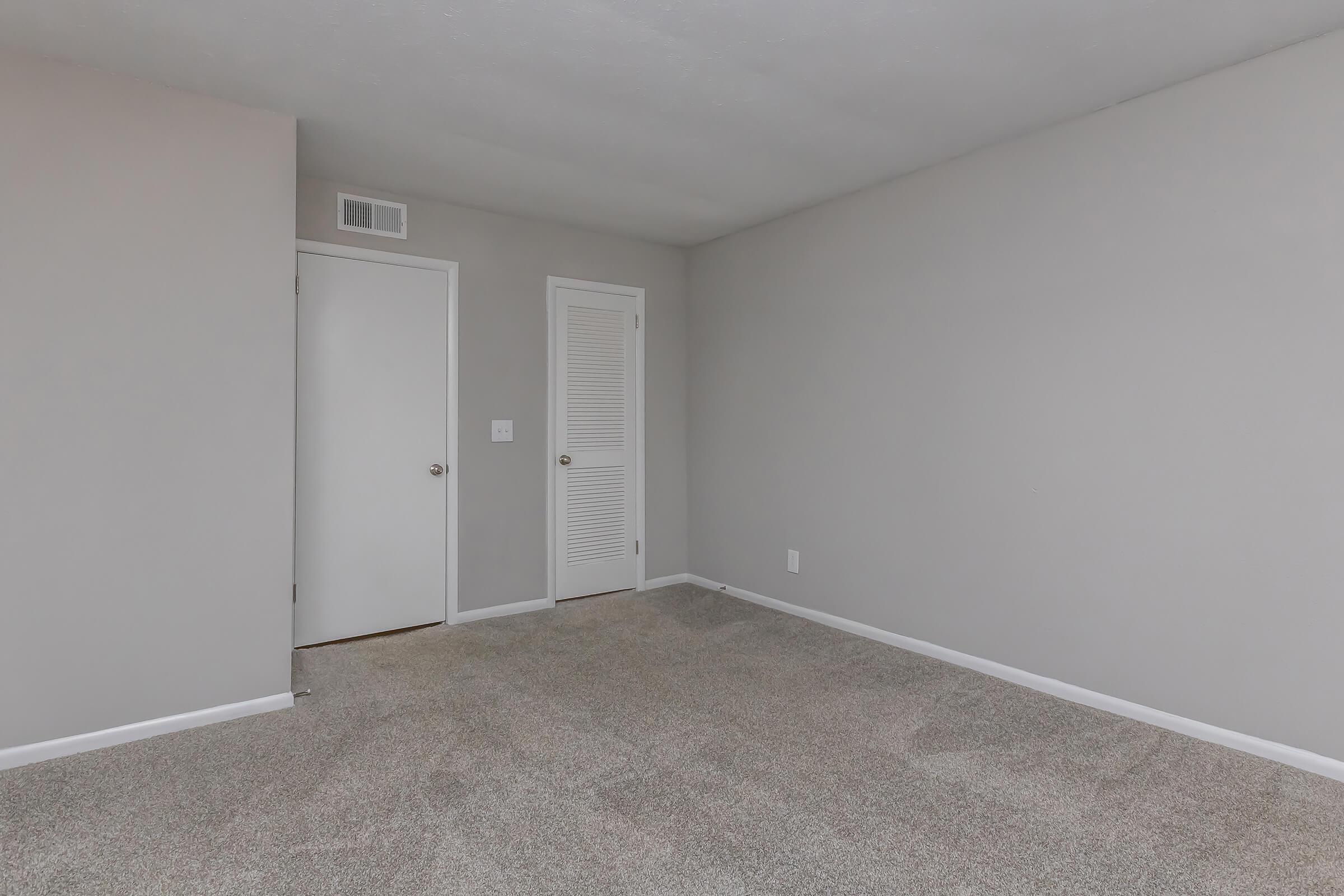 Empty room with light gray walls and beige carpet. Features a closed door on the left side and a closet door on the right. Minimalistic layout with no furniture or decorations. Natural light flooding in from an unseen window, creating a spacious feel.