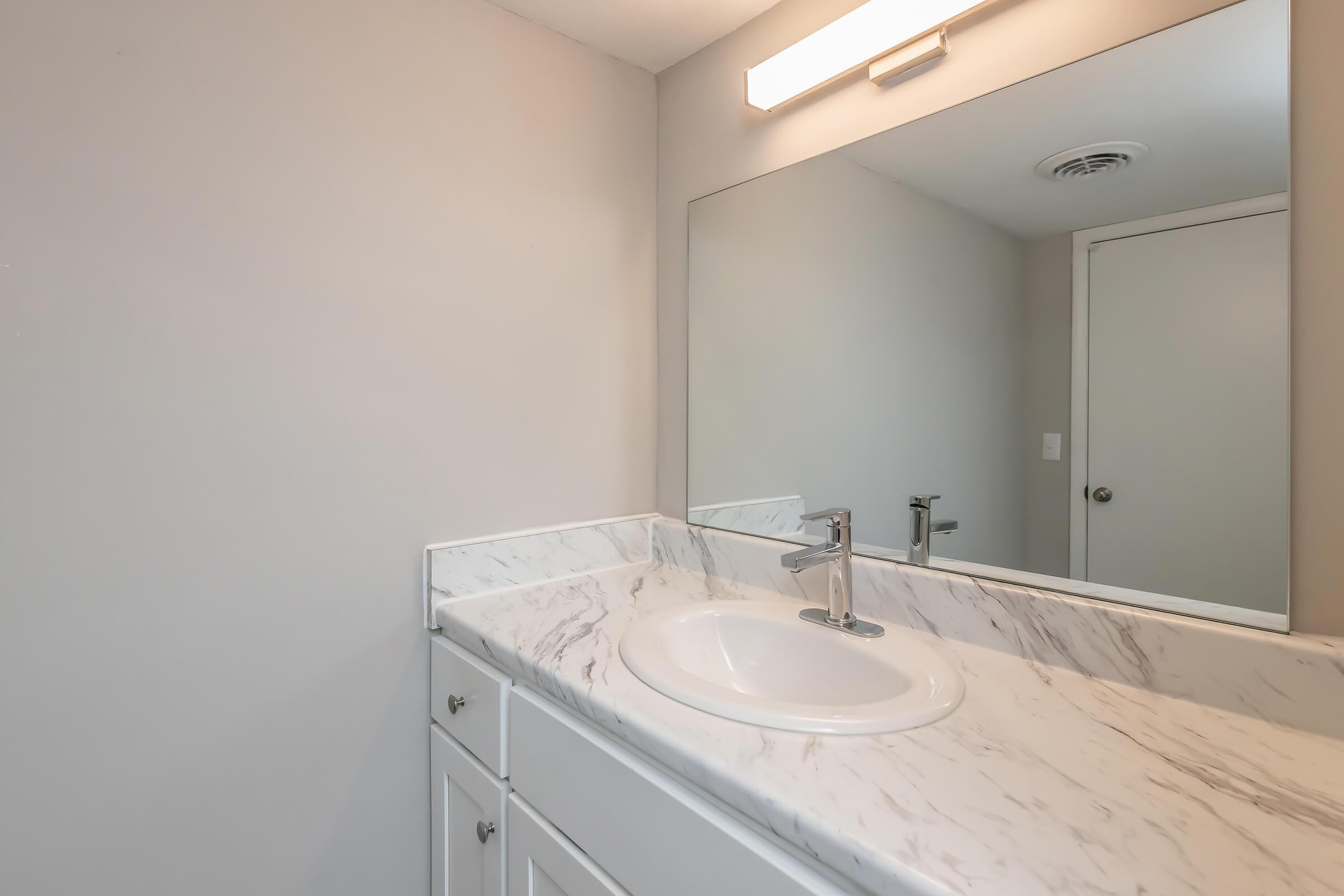 A clean and modern bathroom featuring a white marble countertop with a sink, a large mirror reflecting the room, and soft lighting above. The walls are a light gray color, and a door is visible in the background, adding to the overall minimalist design.