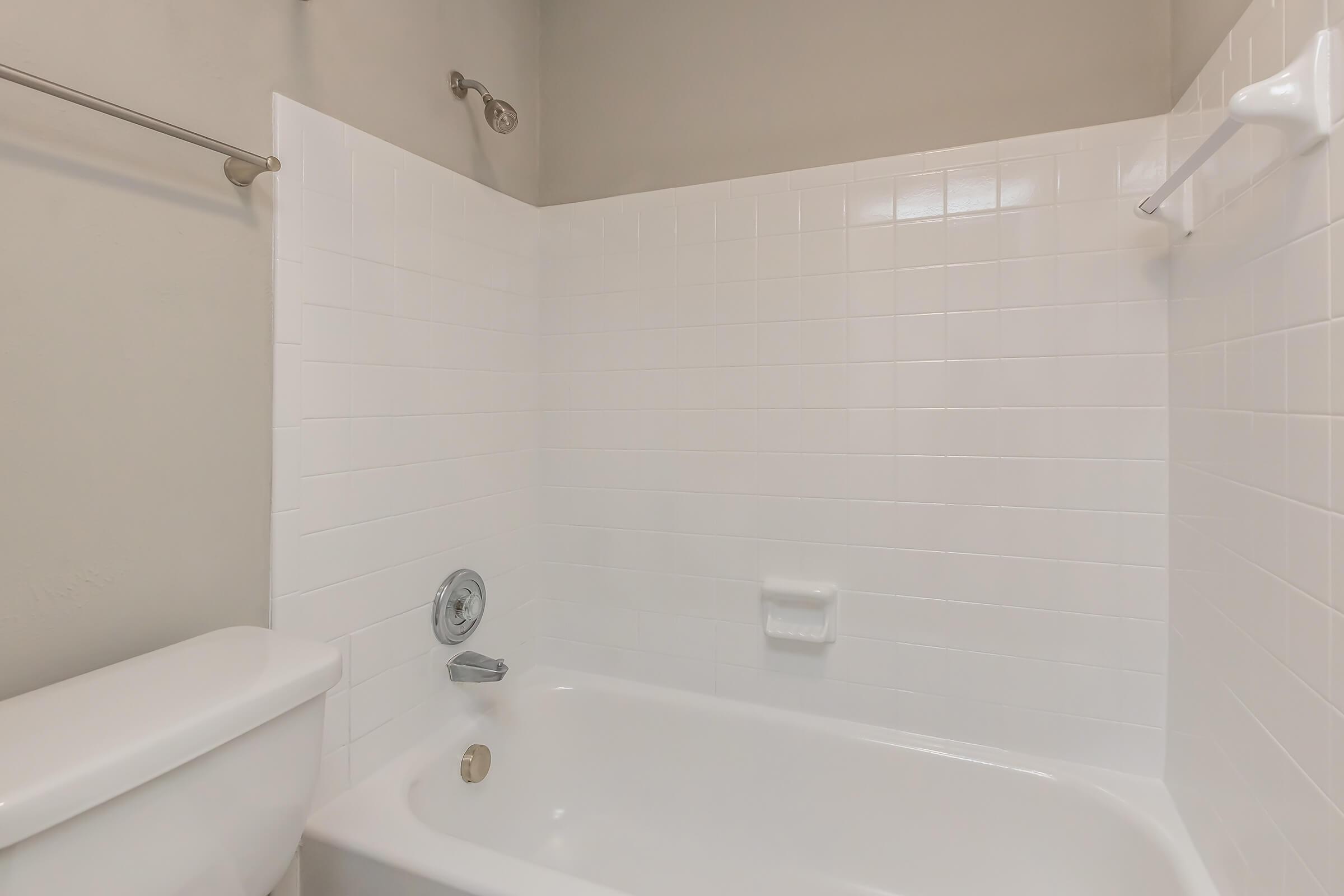 A clean, modern bathroom with a white tiled shower and bathtub. The bathtub features a chrome faucet and a wall-mounted showerhead. There is a white toilet nearby, and the walls are painted a soft neutral color. The overall design is simple and minimalist.