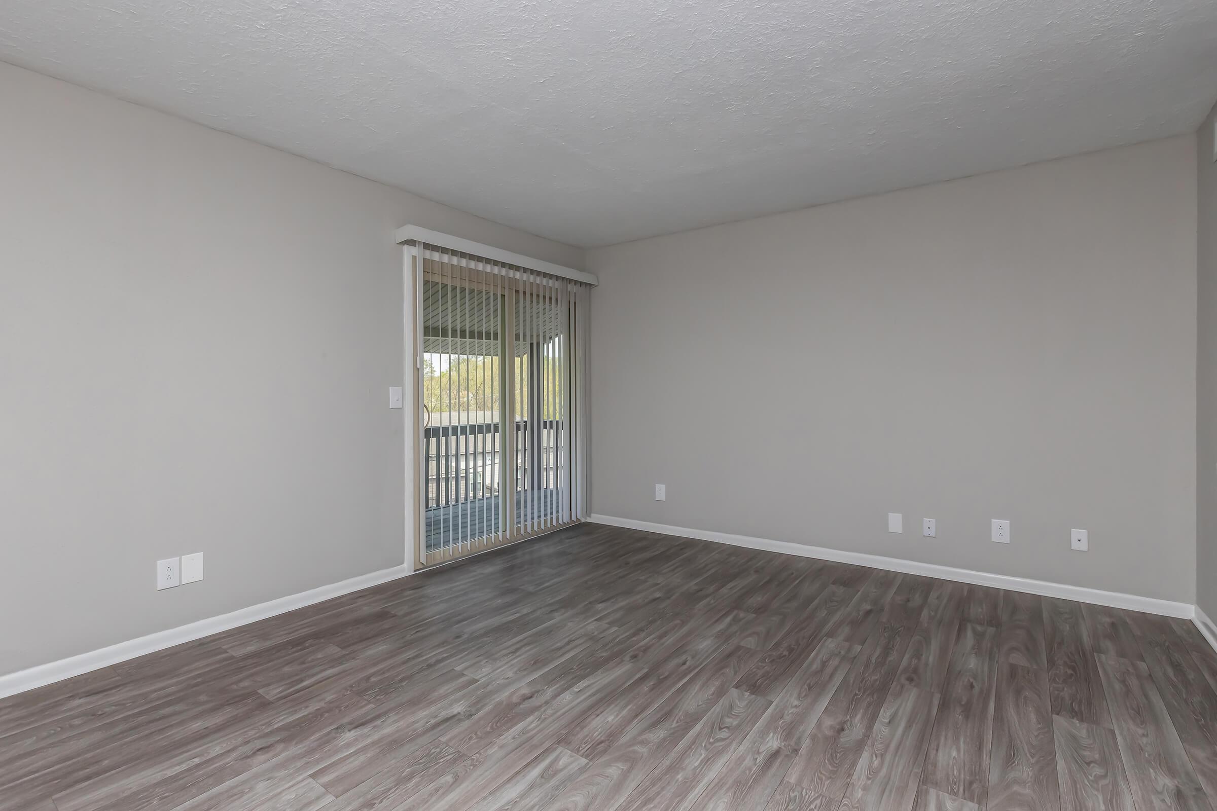 Interior view of a spacious, empty room with light gray walls and wooden flooring. Floor-to-ceiling sliding glass doors lead to a balcony, allowing natural light. The room has minimal decorative elements, creating a blank canvas for potential furnishing and decoration.