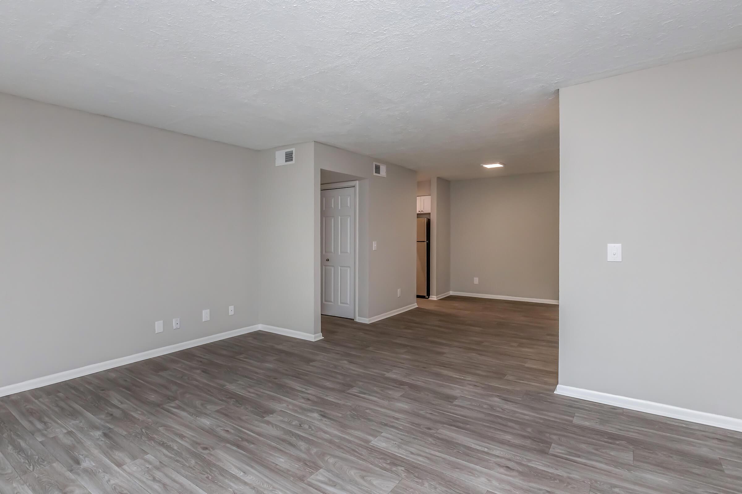Spacious interior of an apartment featuring light-colored walls, a textured ceiling, and a large open area with wood-style flooring. A doorway leads to a kitchen area in the background, and there are several light switches on the walls.