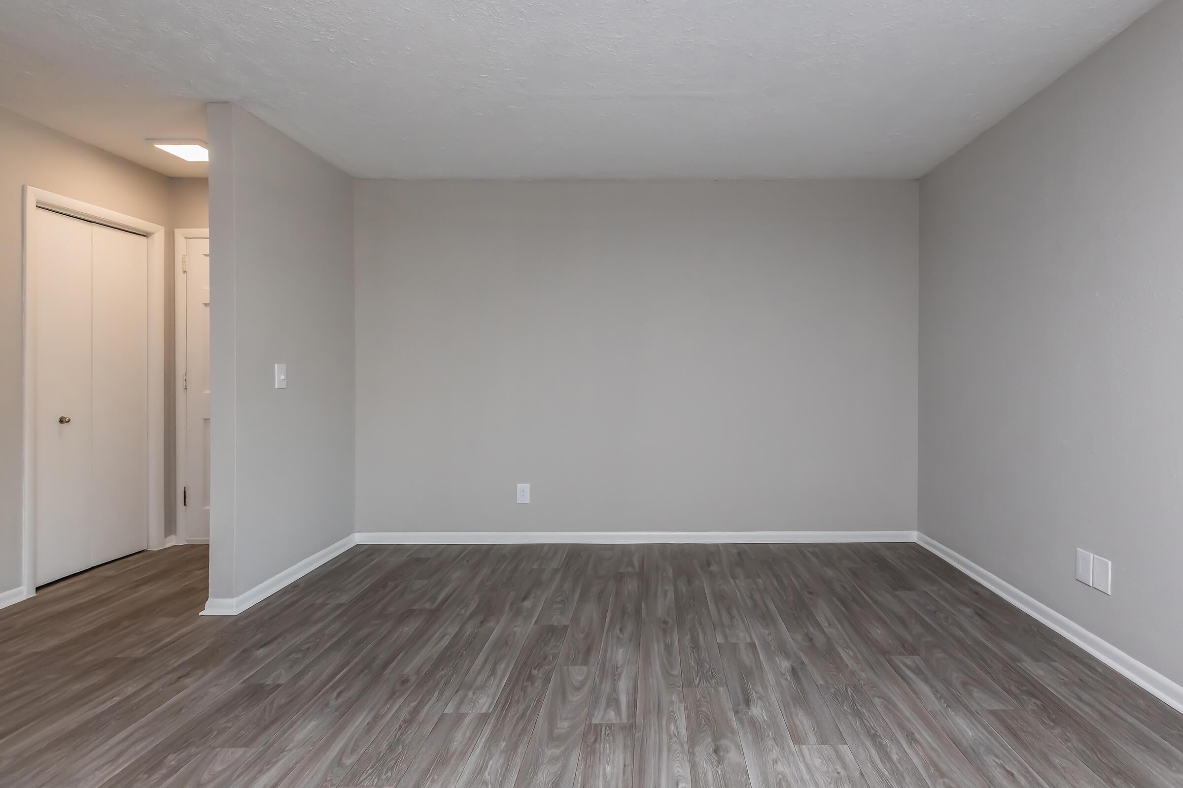 A spacious, empty room with light gray walls and a smooth, light wood-like flooring. A single door leads to another room, with no furniture present, creating an open and airy atmosphere. Natural light enters from a small window, enhancing the sense of space.