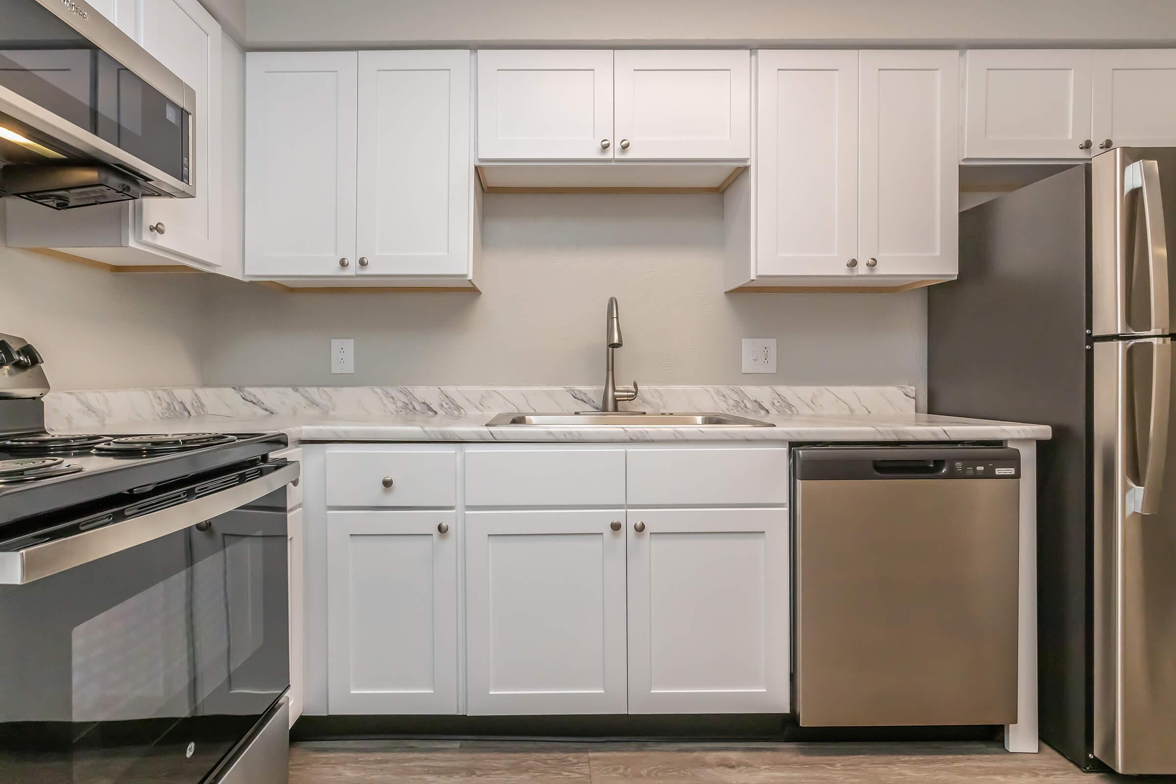 Modern kitchen featuring white cabinetry with silver hardware, a stainless steel refrigerator, a dishwasher, and a stove. The countertop is made of light-colored marble, and there's a sleek sink with a pull-down faucet. The overall design is clean and contemporary, with a focus on functionality.